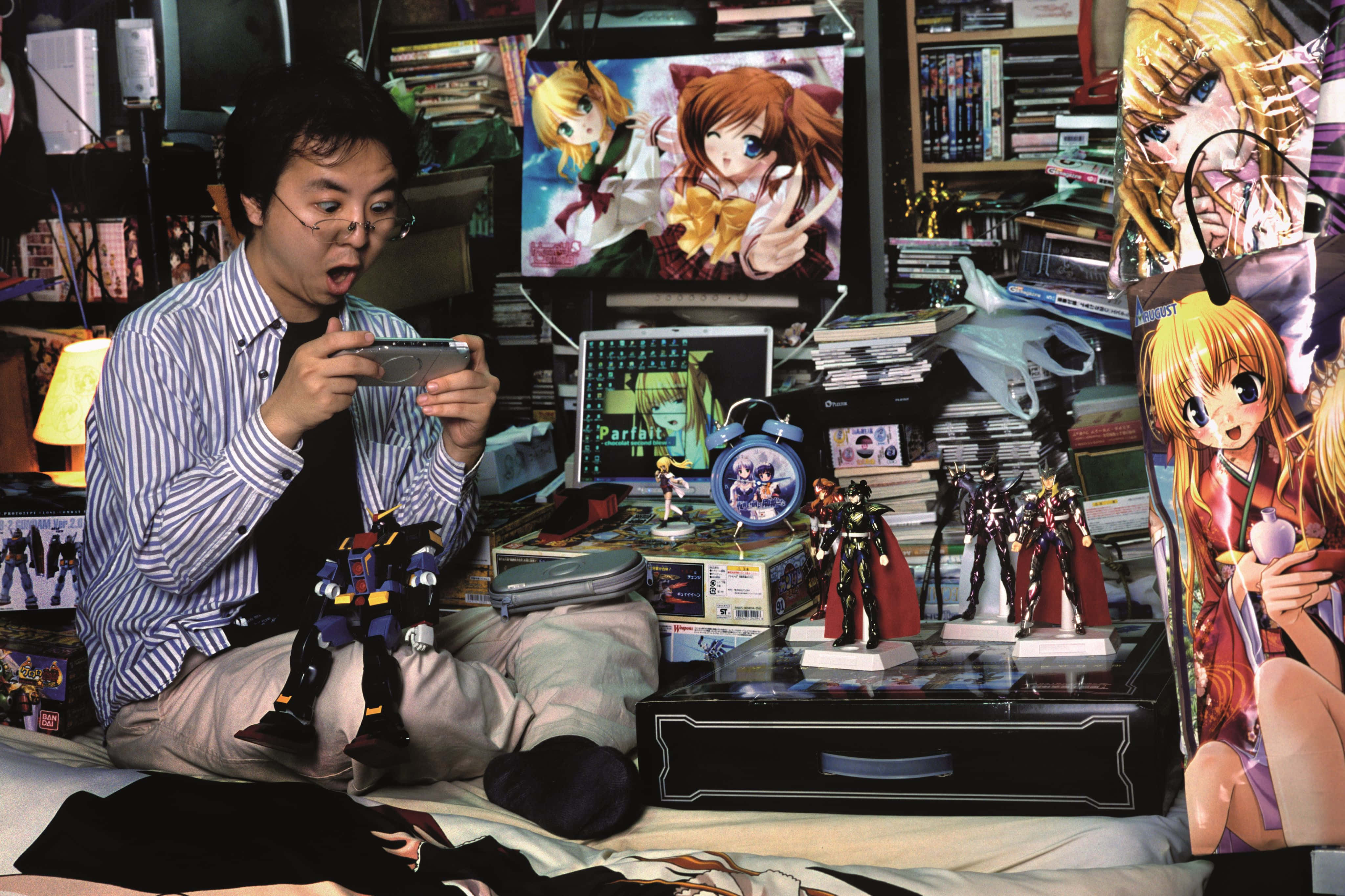 Otaku 桌布 壁紙 — 一個男人坐在床上，周圍是動漫海報、公仔和遊戲裝備，專注於他的手持裝置。