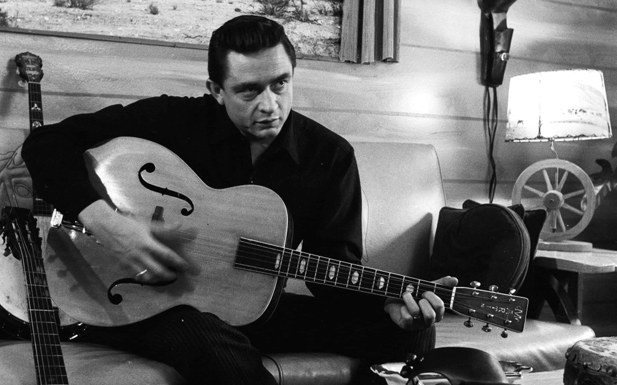johnny cash 桌布 壁紙 — 一個男人坐在沙發上，拿著一把空心吉他，在一個溫暖燈光的復古房間裡，有木牆和一盞燈。