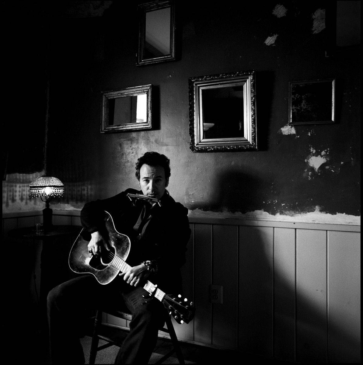 Bruce Springsteen 桌布 壁紙 — 一個男人坐在燈光昏暗的房間裡，拿著吉他和口琴，身後有復古鏡子和剝落的壁紙。