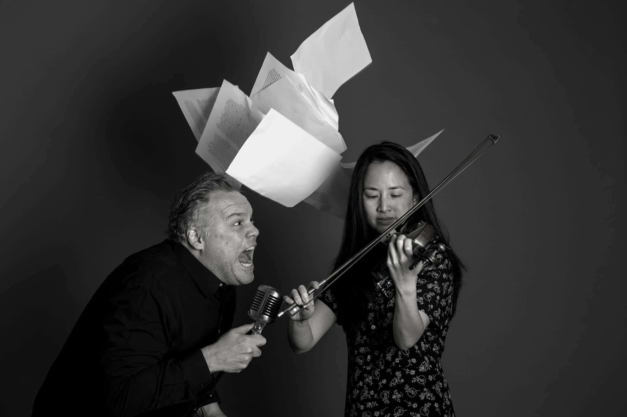 Vincent D'onofrio 桌布 壁紙 — 一個男人對著復古麥克風尖叫，紙張在一個拉小提琴的女人上方爆炸，場景在鮮明的黑白攝影棚中。