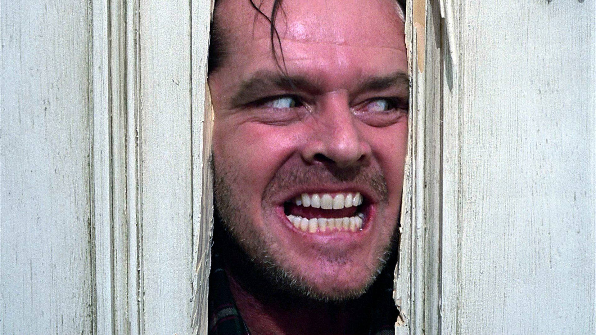 The Shining 桌布 壁紙 — 一個男人帶著瘋狂的笑容，透過破爛的白色木門框，用銳利的眼神窺視。