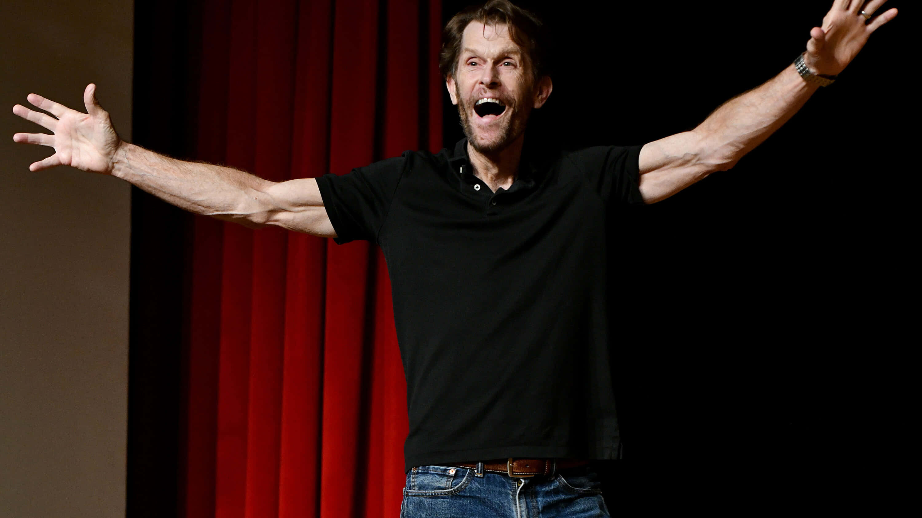 Kevin Conroy 桌布 壁紙 — 一個男人張開雙臂，張開嘴巴，發出喜悅的驚嘆，站在紅色窗簾前，背景黑暗。