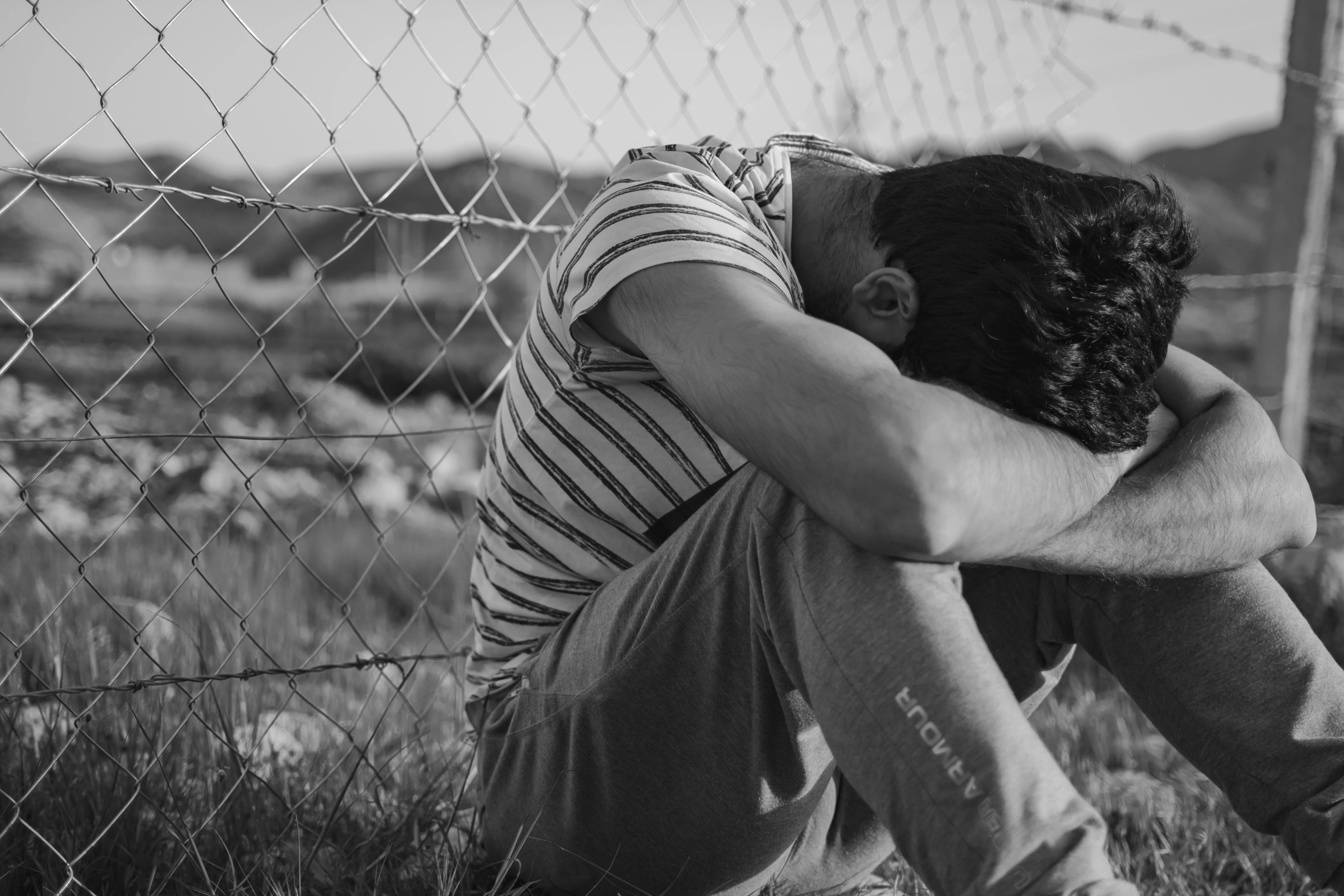 crying sad boy 桌布 壁紙 — 一個男人彎腰悲傷地坐在荒涼田野的鐵絲網圍欄旁，雙臂環抱膝蓋，黑白照片。