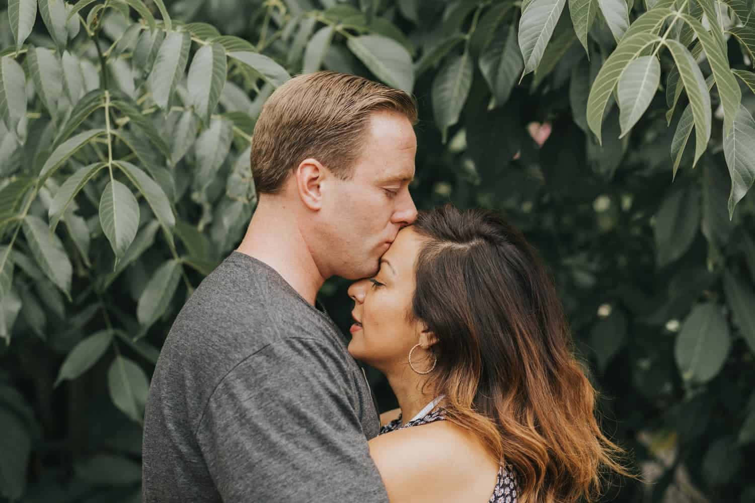 couple love 桌布 壁紙 — 一個男人溫柔地親吻一個女人的額頭，他們在茂密的綠色樹葉下擁抱，一個溫柔的時刻。