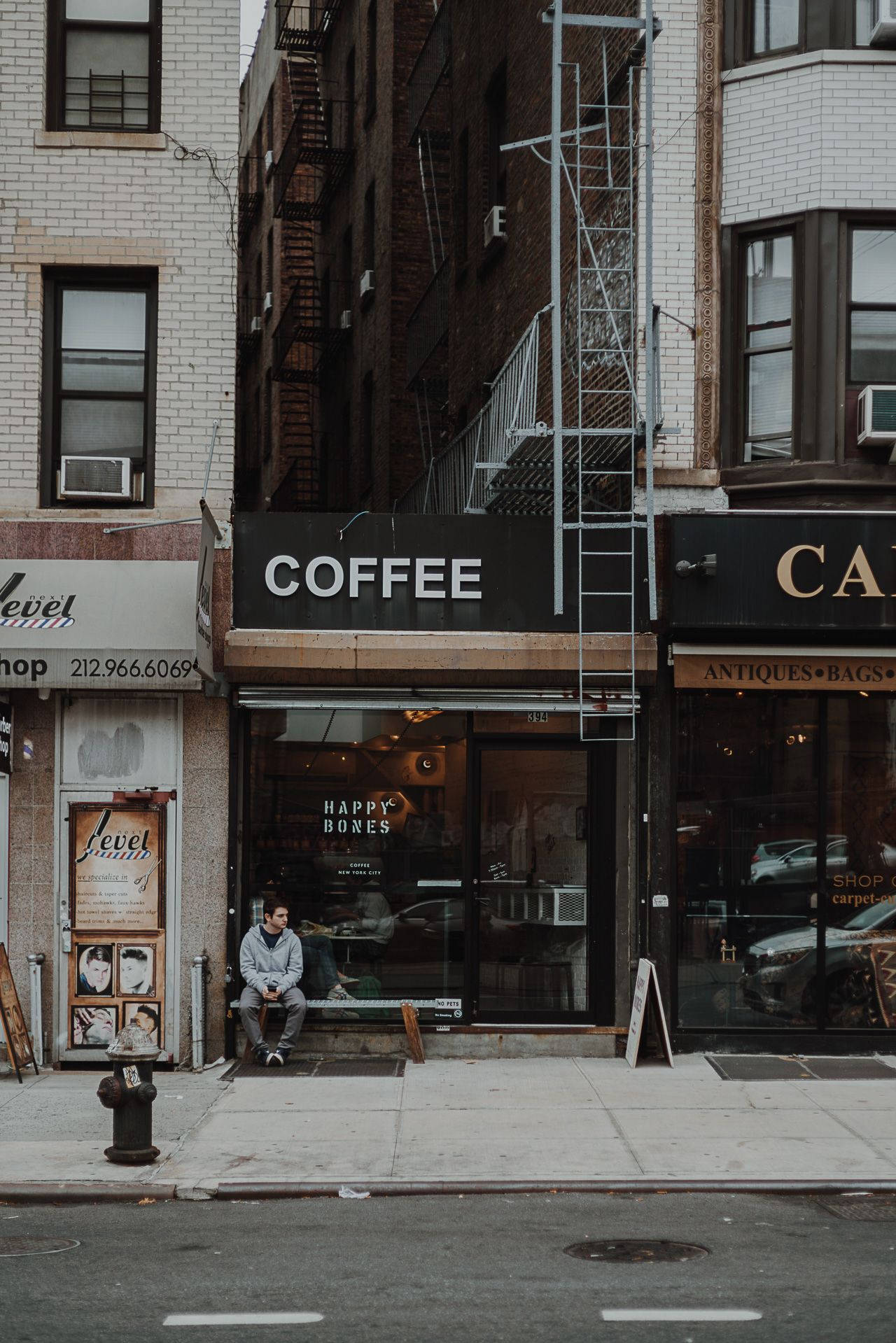 New York 桌布 壁紙 — 一個男人獨自坐在城市街道上咖啡店外的人行道長椅上，周圍有消防逃生梯和磚砌建築。