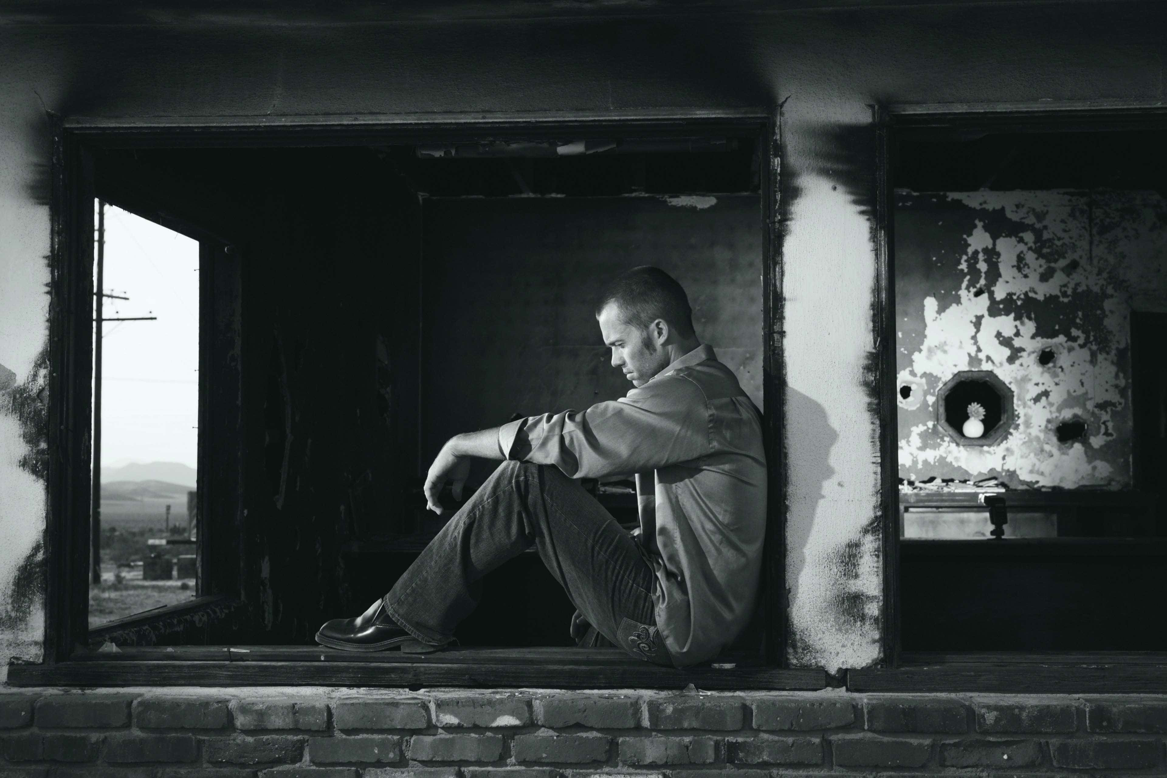Depressing 桌布 壁紙 — 一個男人獨自坐在燒毀的房間裡，透過窗戶凝視遠方的風景，呈現強烈的黑白對比。
