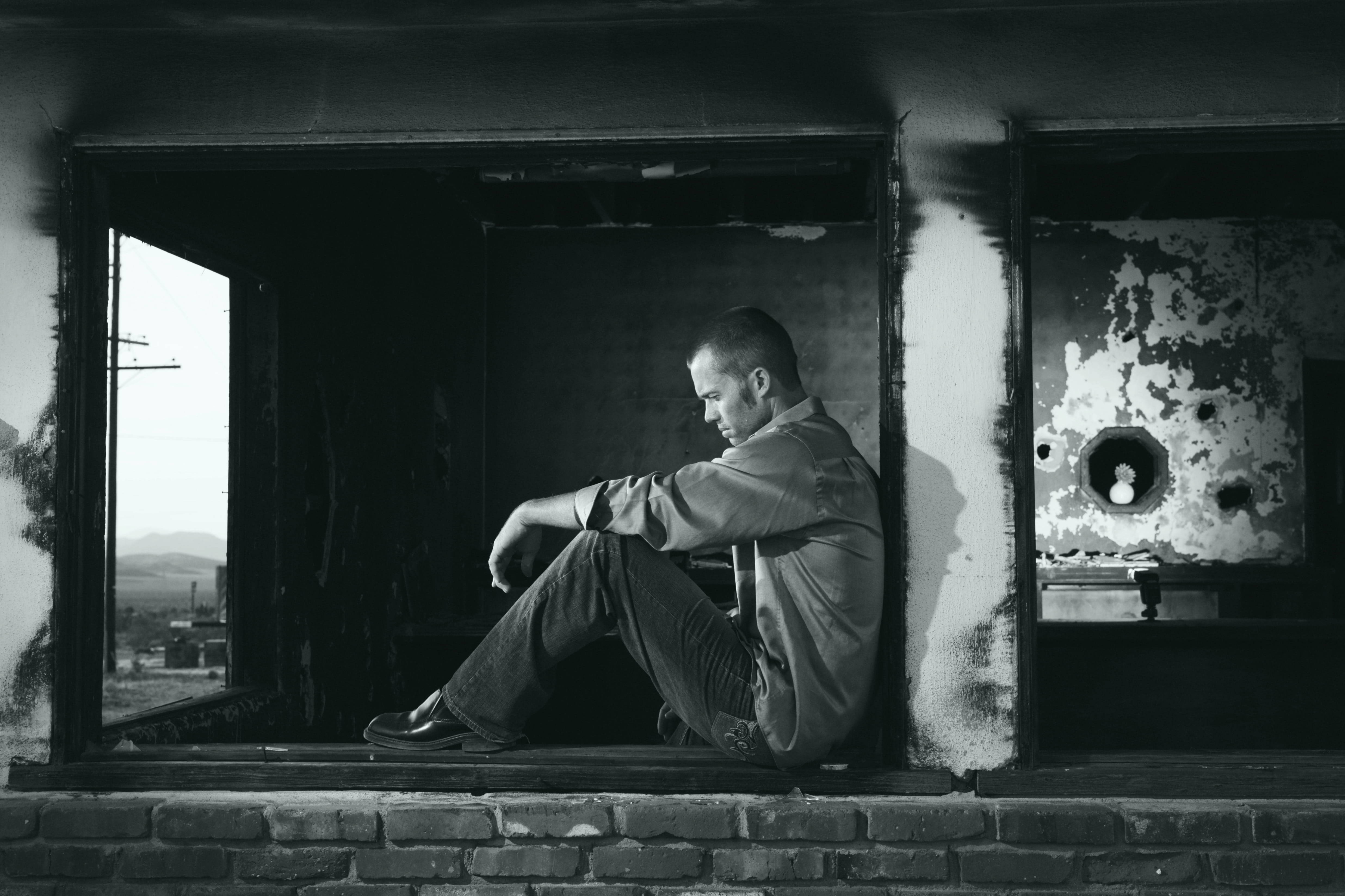 Sitting Alone 桌布 壁紙 — 一個男人獨自坐在破舊的房間裡，透過破碎的窗戶凝視遠方的風景，呈現強烈的黑白對比。