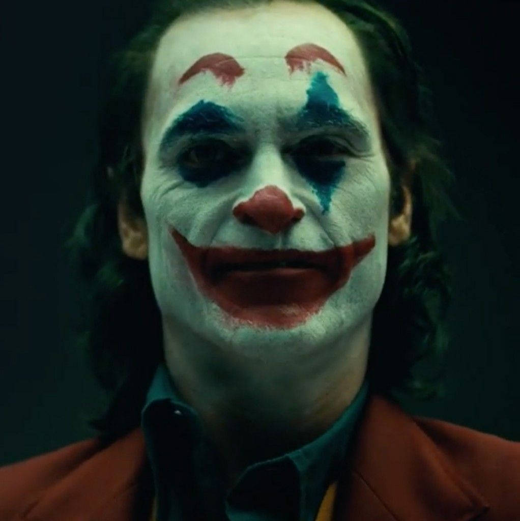 Joker 2019 桌布 壁紙 — 一個男人的特寫，臉上塗著白色顏料，紅唇和藍色淚痕，在黑暗背景下強烈凝視。