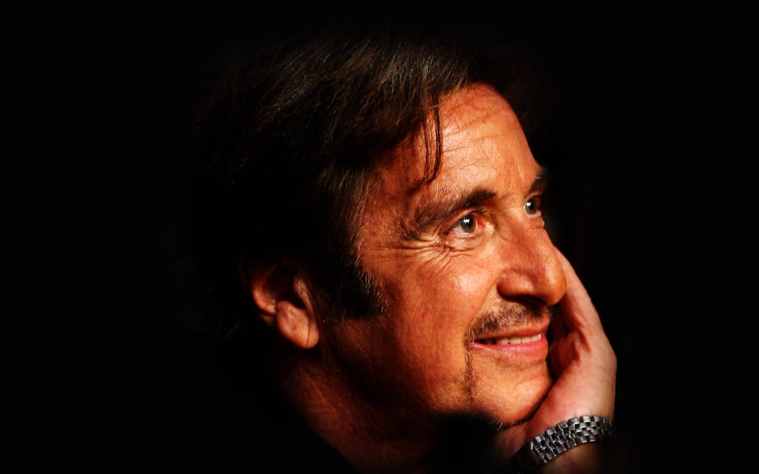 Al Pacino 桌布 壁紙 — 一個男人的特寫肖像，他帶著溫暖的笑容，將下巴托在手上，背景是黑色。