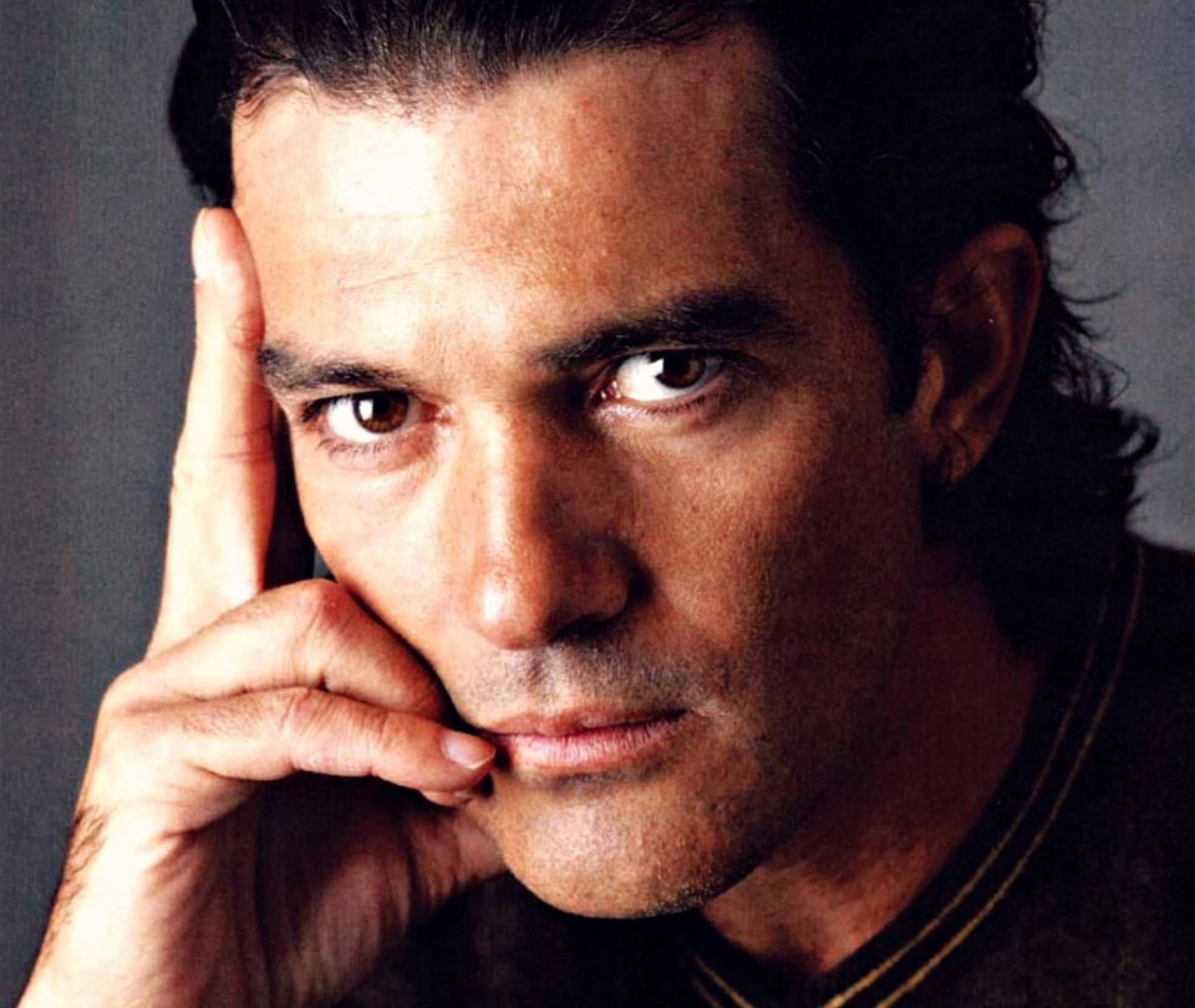 antonio banderas 桌布 壁紙 — 一個男人的特寫肖像，他有強烈的棕色眼睛，將手放在臉頰上，背景是柔和的灰色。