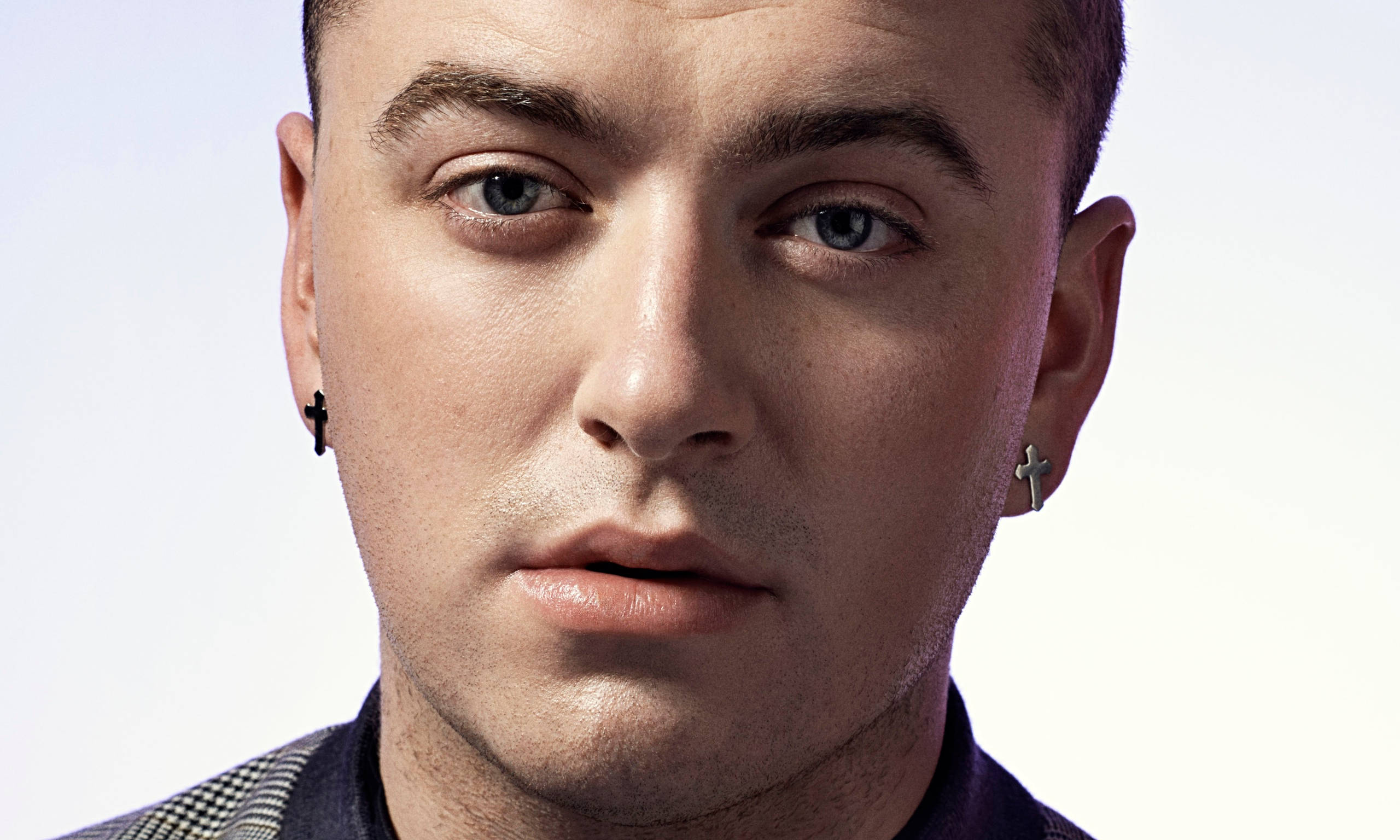 Sam Smith 桌布 壁紙 — 一個男人的特寫肖像，他有強烈的藍色眼睛和十字架耳環，背景是柔和的白色。