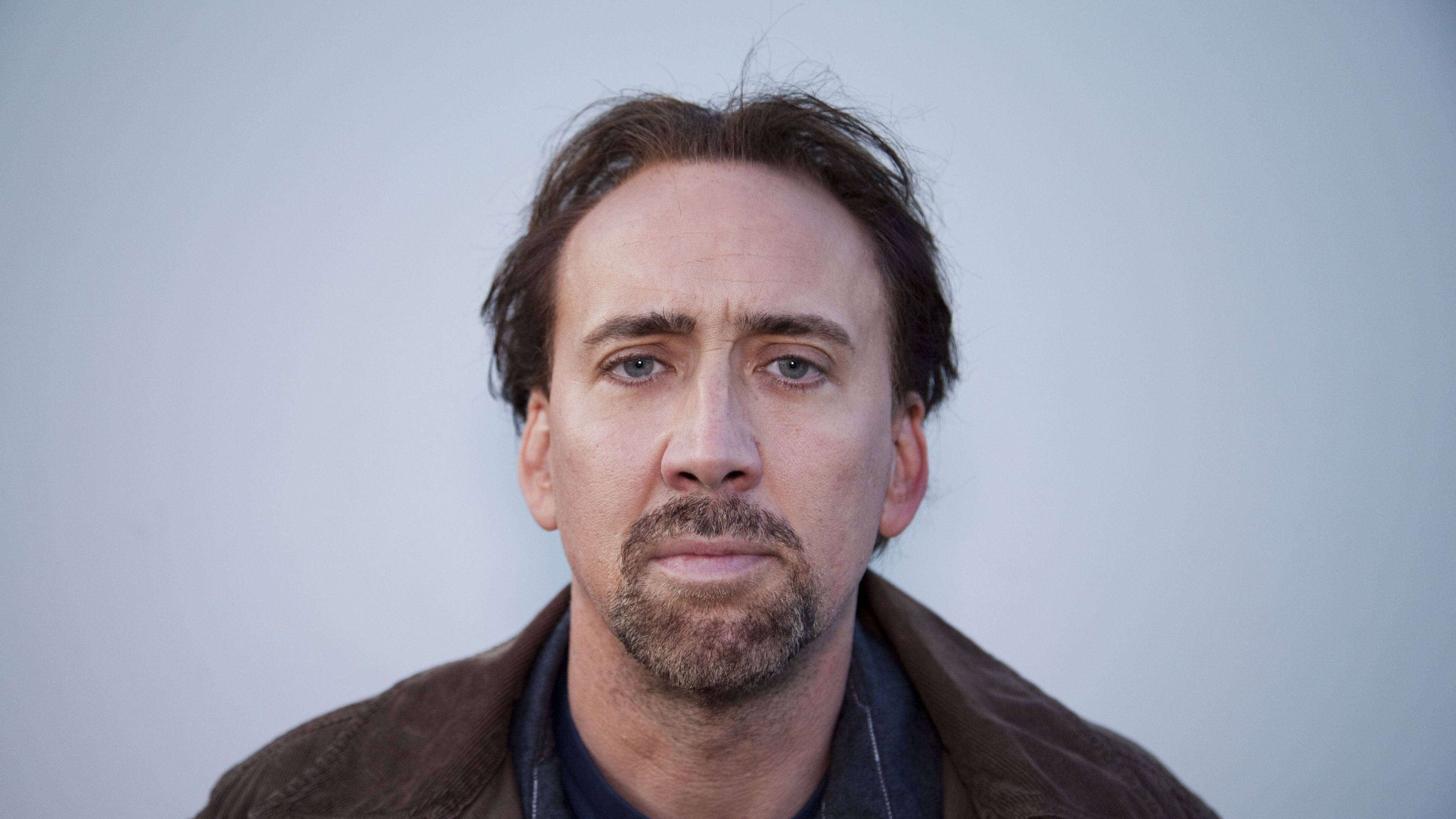 Nicolas Cage 桌布 壁紙 — 一個男人的特寫肖像，表情嚴肅，身穿棕色外套，背景是純淺灰色。