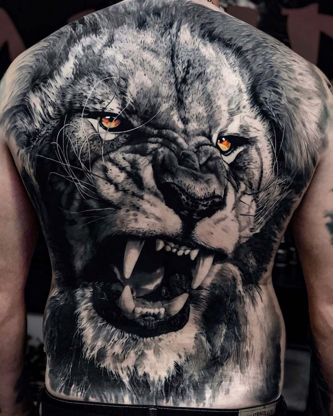 Lion Head 桌布 壁紙 — 一個男人的背部，上面有一個超寫實的黑灰紋身，是一隻咆哮的獅子，有著發光的琥珀色眼睛。