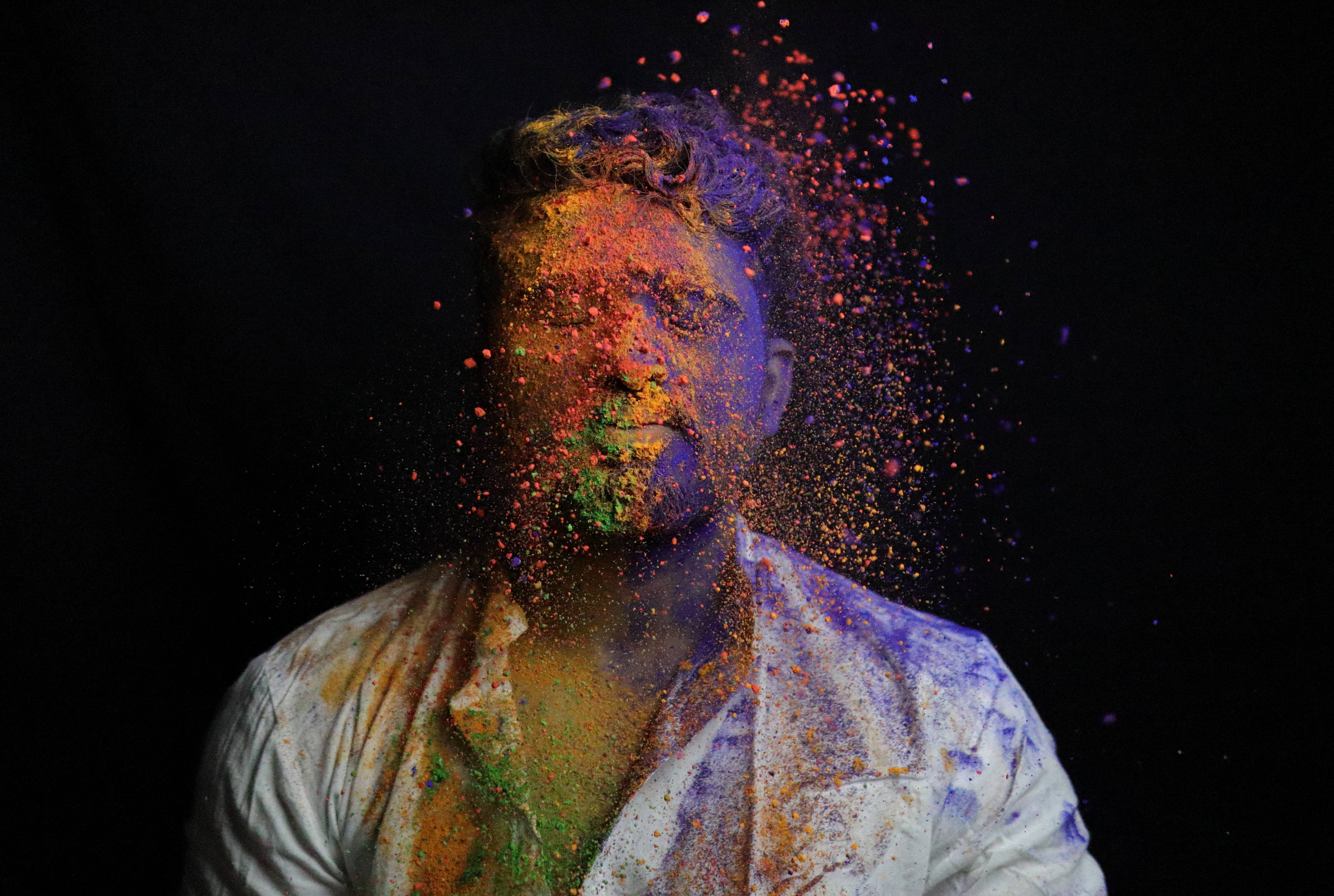 Holi 桌布 壁紙 — 一個男人的臉和襯衫在黑色背景上爆發出鮮豔的彩色粉末。