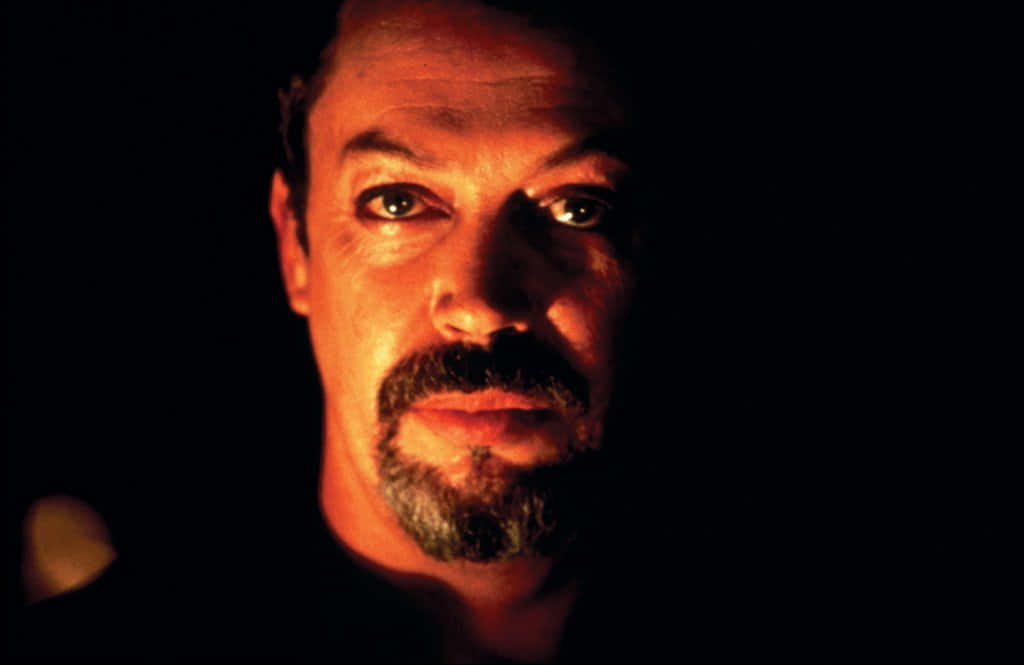 Tim Curry 桌布 壁紙 — 一個男人的臉被溫暖的橙色光線戲劇性地照亮，背景是黑色，他銳利的目光穿透陰影。
