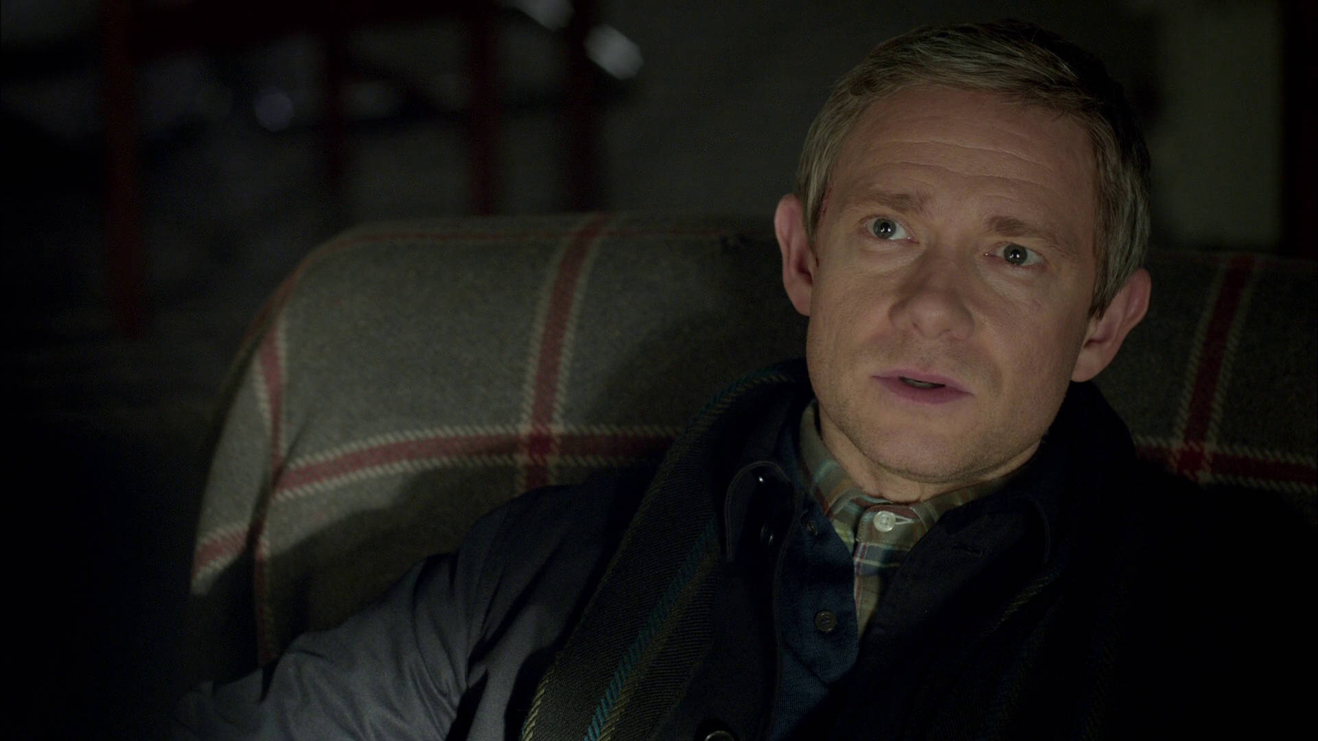 Martin Freeman 桌布 壁紙 — 一個男人睜大眼睛坐在昏暗房間的格紋沙發上，看起來很吃驚。
