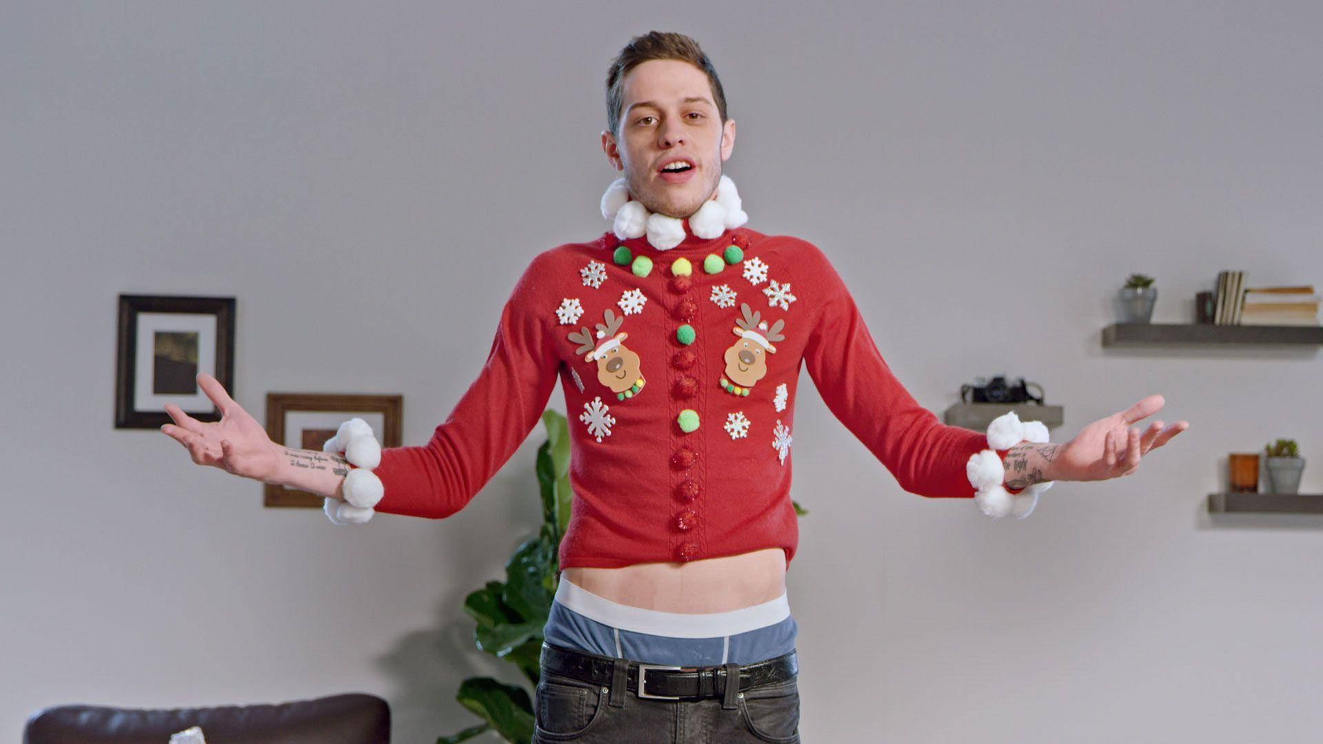 Pete Davidson 桌布 壁紙 — 一個男人穿著紅色醜陋的聖誕毛衣，上面有雪花、馴鹿和絨球，站在簡約的客廳裡，張開雙臂做手勢。