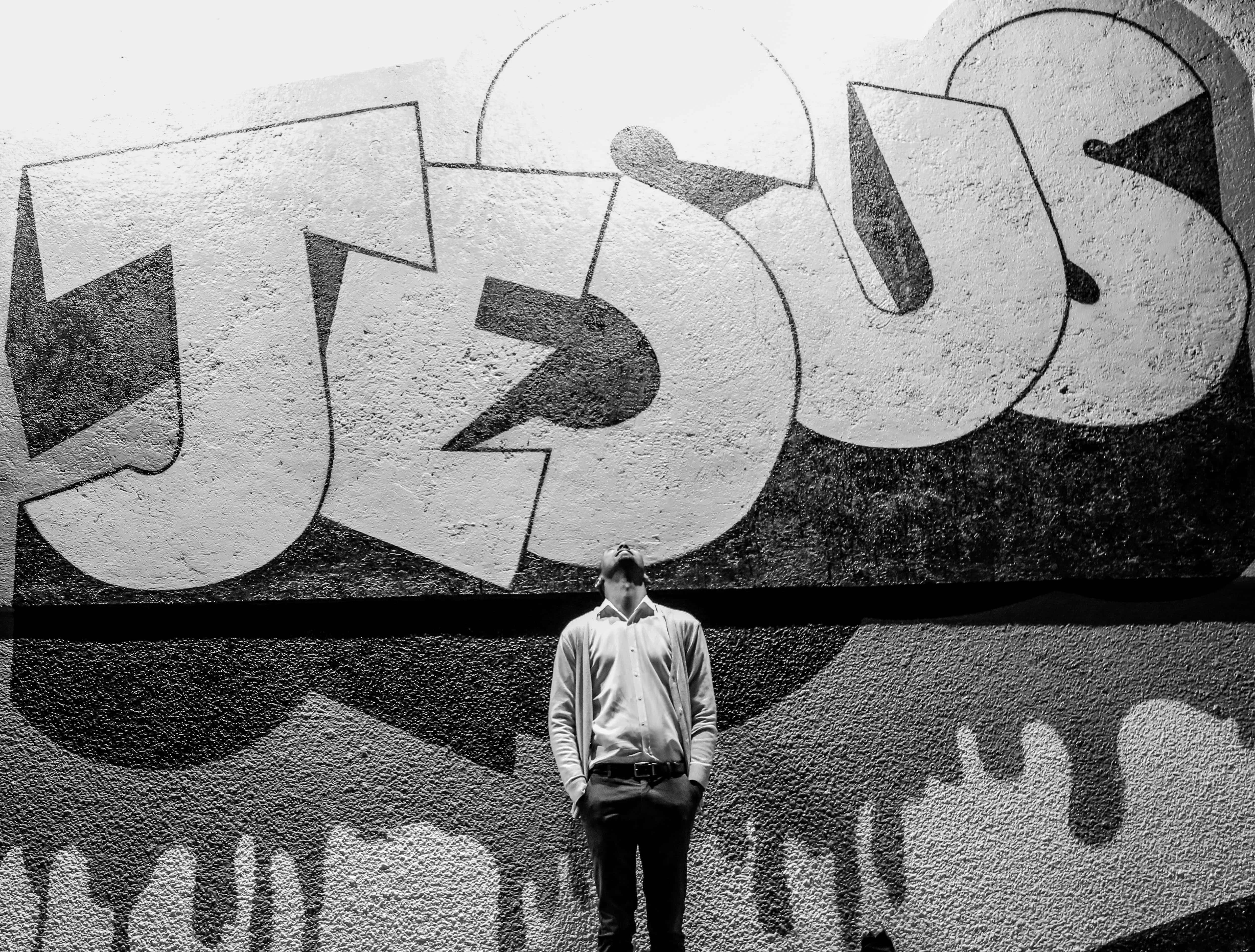 Jesus iPhone 桌布 壁紙 — 一個男人站在一幅巨大的黑白塗鴉壁畫前，壁畫在紋理牆上拼出 JESUS。