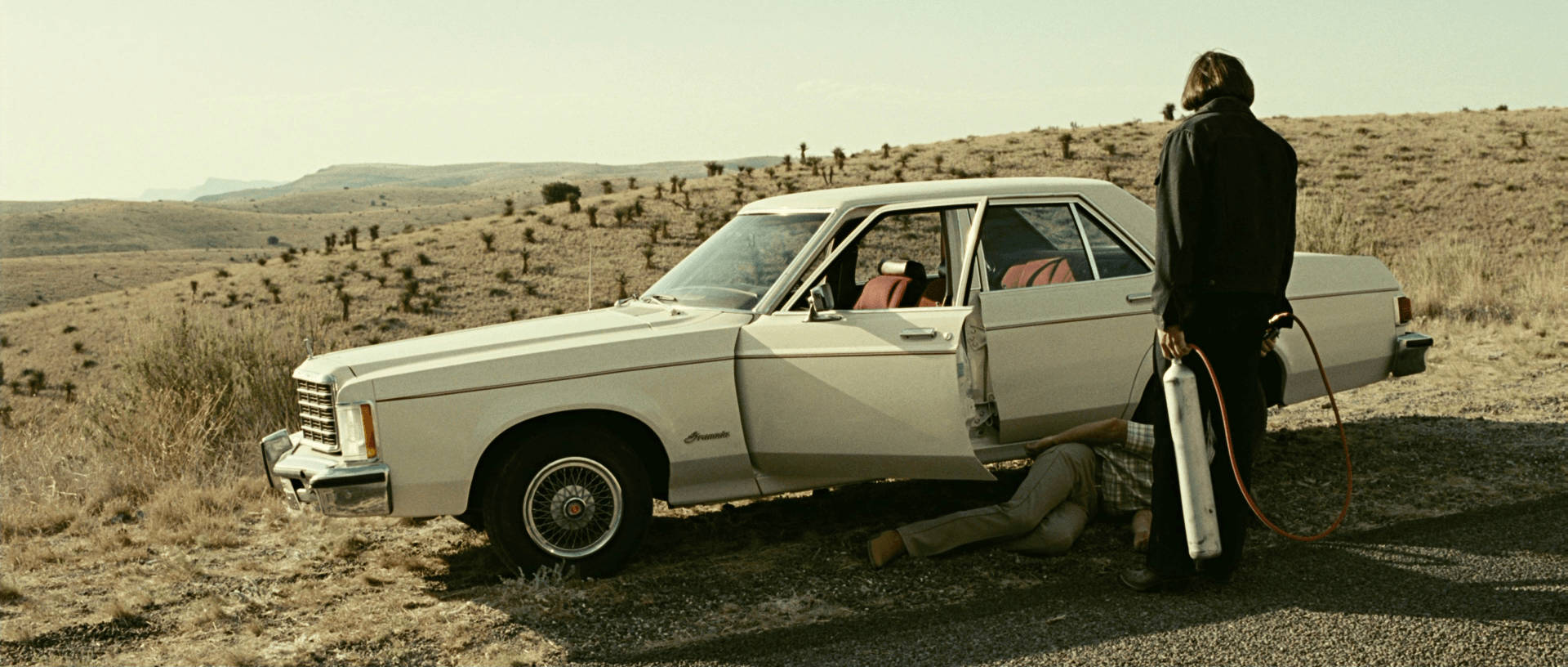 Javier Bardem 桌布 壁紙 — 一個男人站在一輛白色復古車旁邊，在乾燥的沙漠景觀中，另一個人躺在附近的地上。