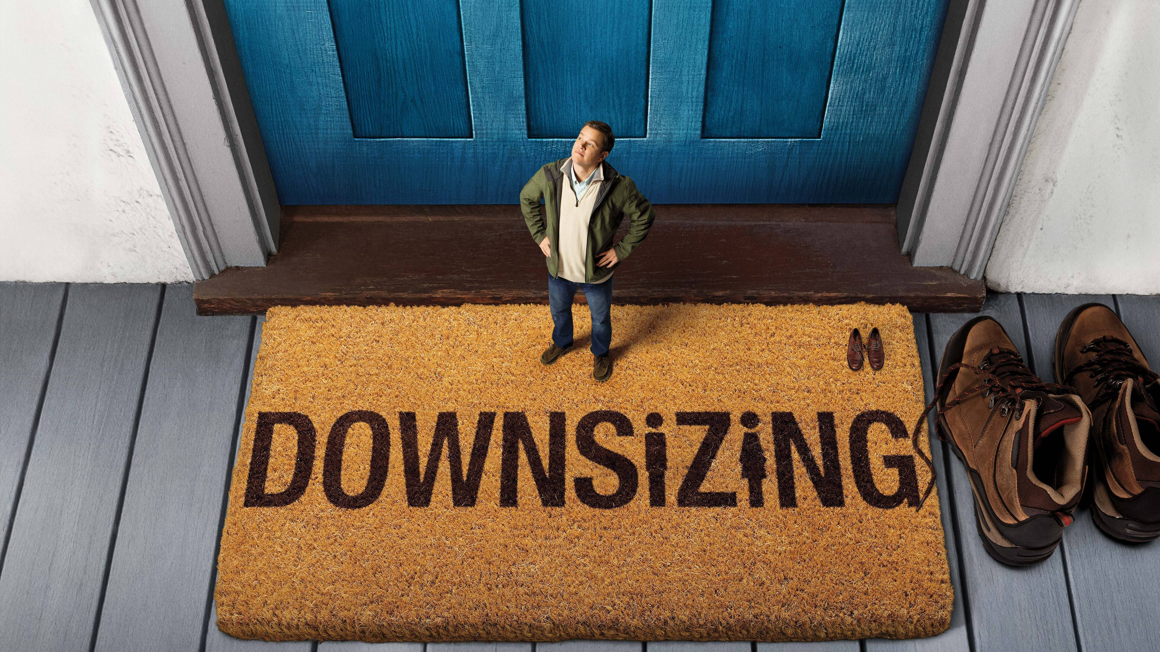 麥特·戴蒙 桌布 壁紙 — 一個男人站在寫著「DOWNSIZING」的門墊上，旁邊有一扇藍色門，鞋子整齊地放在一旁。