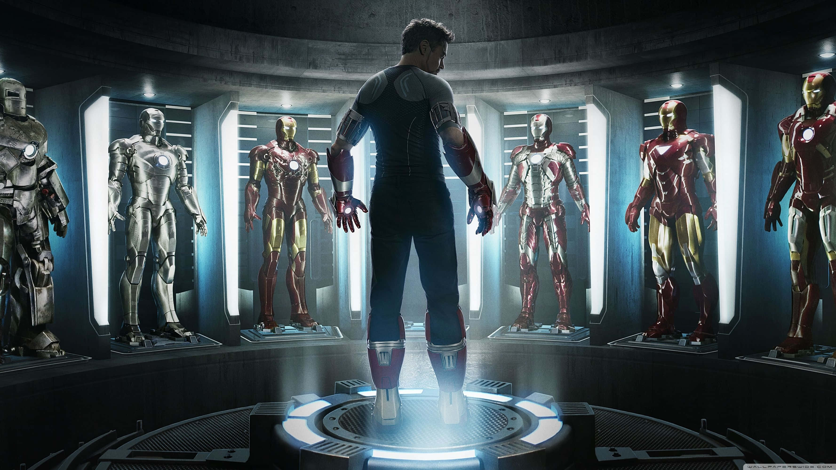 iron man 3 桌布 壁紙 — 一個男人站在發光平台上，面向一個圓形顯示器，展示著各種鋼鐵人戰衣，位於高科技實驗室中。