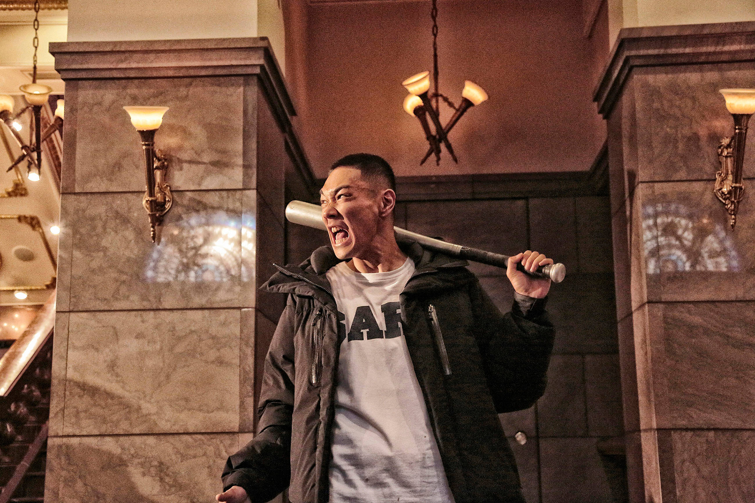prisoners of the ghostland 桌布 壁紙 — 一個男人肩上扛著棒球棍，在一個由復古壁燈照亮的宏偉大理石走廊中尖叫。