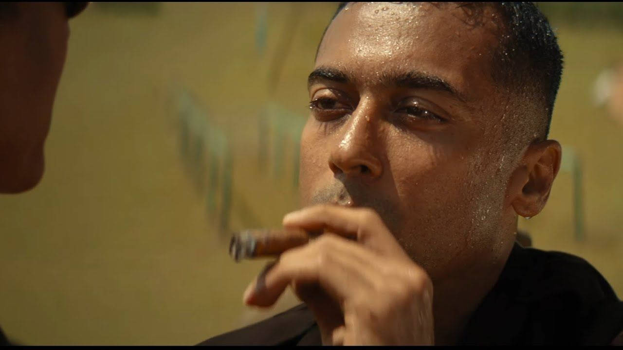 Indian 桌布 壁紙 — 一個男人臉上閃爍著汗水，在昏暗的房間裡，身後有模糊的綠色圖案，他將雪茄靠近嘴唇。