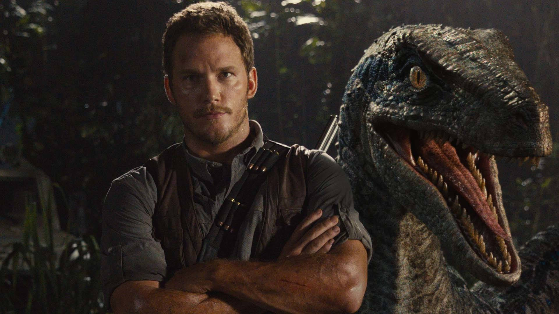Chris Pratt 桌布 壁紙 — 一個男人自信地交叉雙臂，站在夜間茂密叢林中咆哮的猛禽旁邊。