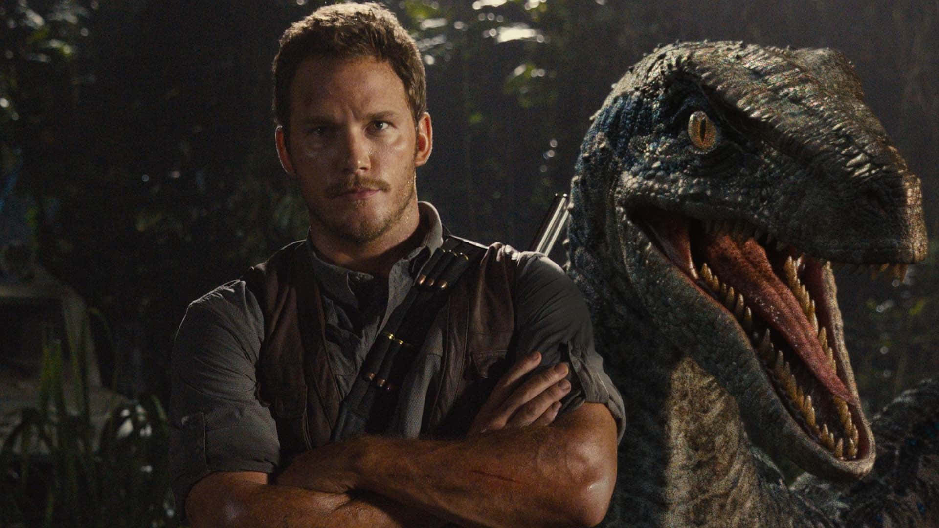 Jurassic World 桌布 壁紙 — 一個男人自信地交叉雙臂，站在昏暗叢林場景中咆哮的迅猛龍旁邊。