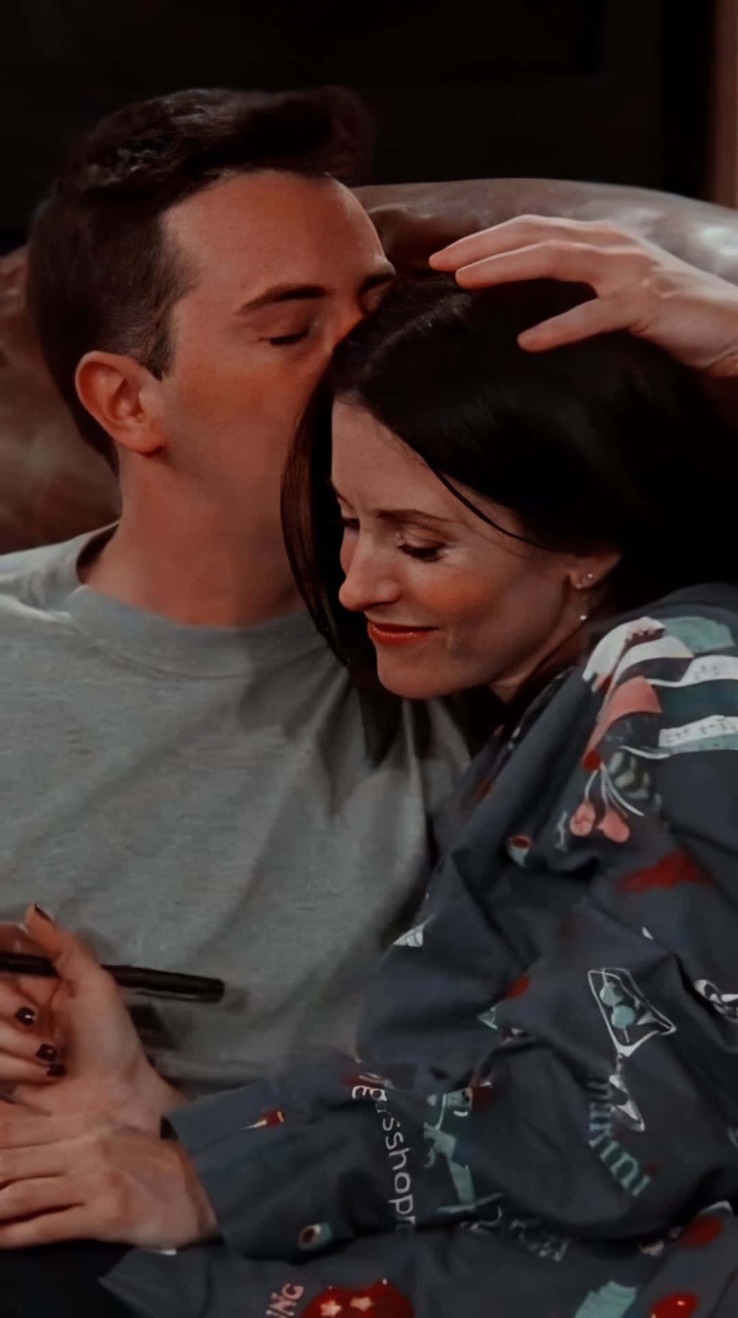 show 桌布 壁紙 — 一個男人親吻一個女人的額頭，而她坐在皮沙發上溫柔地微笑。
