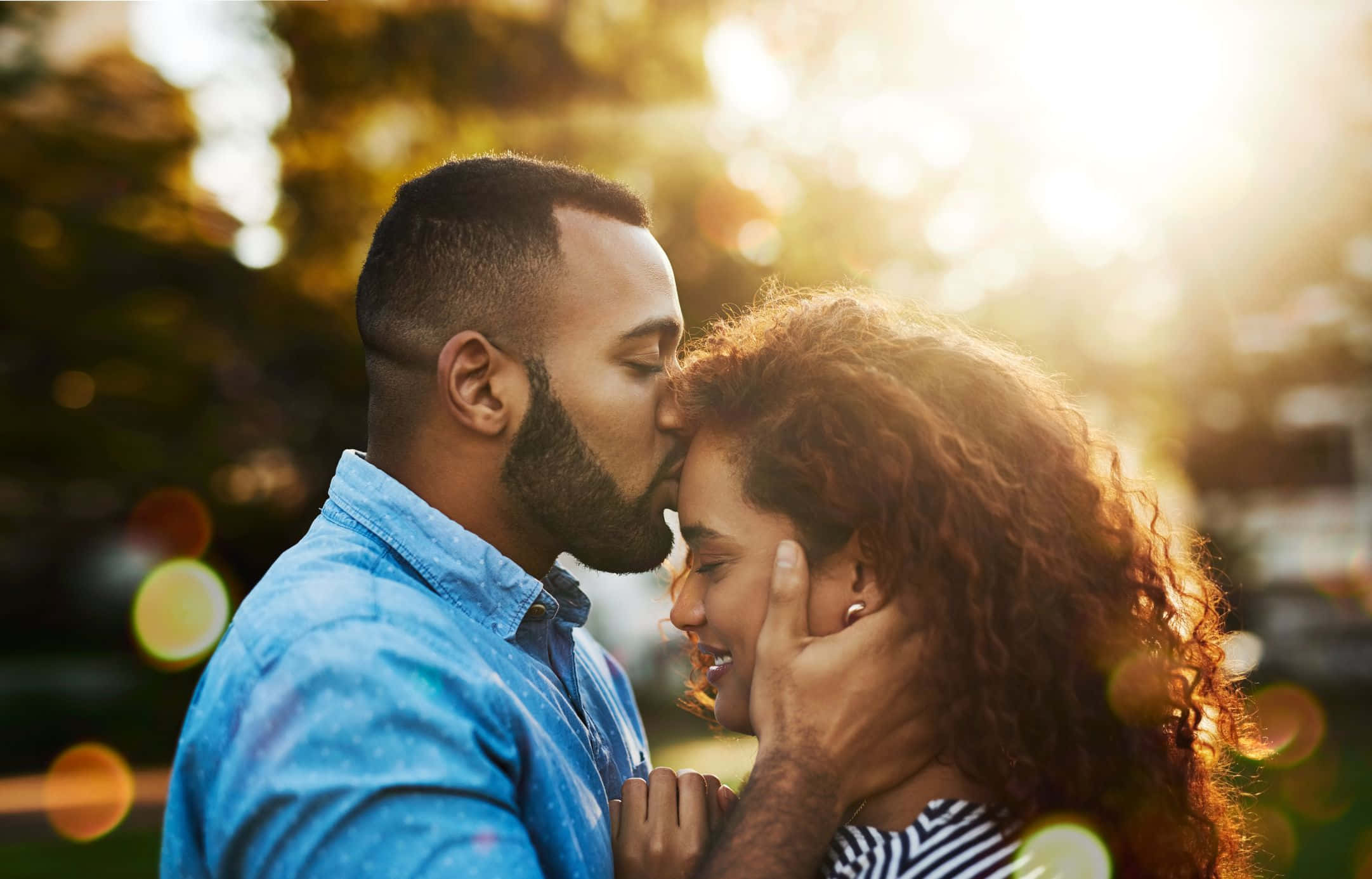 Forehead Kiss 桌布 壁紙 — 一個男人親吻一個微笑女人的額頭，金色的陽光透過他們身後的樹木灑落。