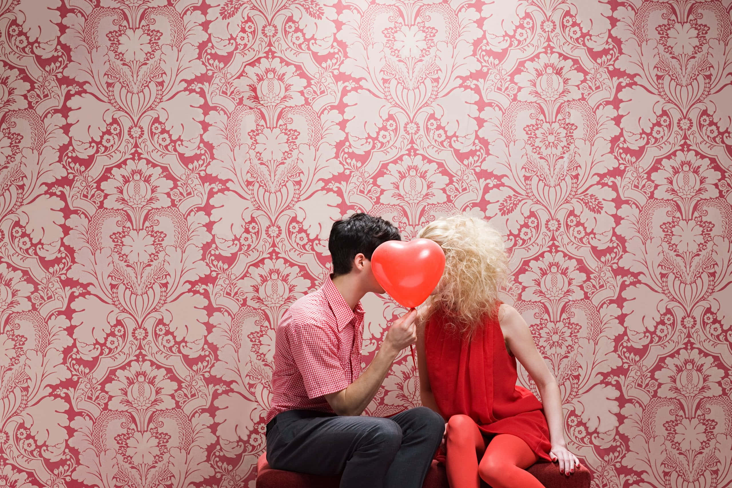 Falling In Love 桌布 壁紙 — 一個男人親吻一個紅色心形氣球，氣球放在一個女人面前，背景是華麗的粉紅色壁紙。