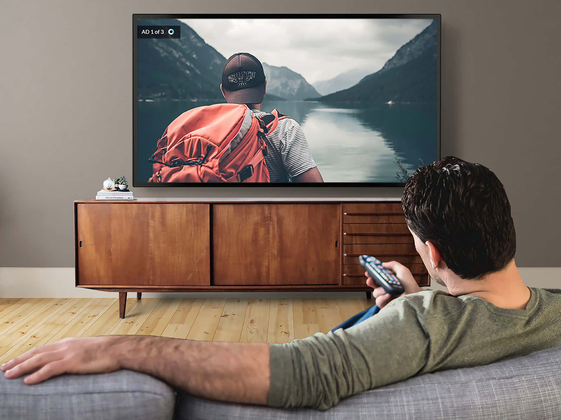 watching tv 桌布 壁紙 — 一個男人躺在沙發上拿著遙控器，看著電視螢幕顯示一個背著紅色背包的遠足者俯瞰山湖。