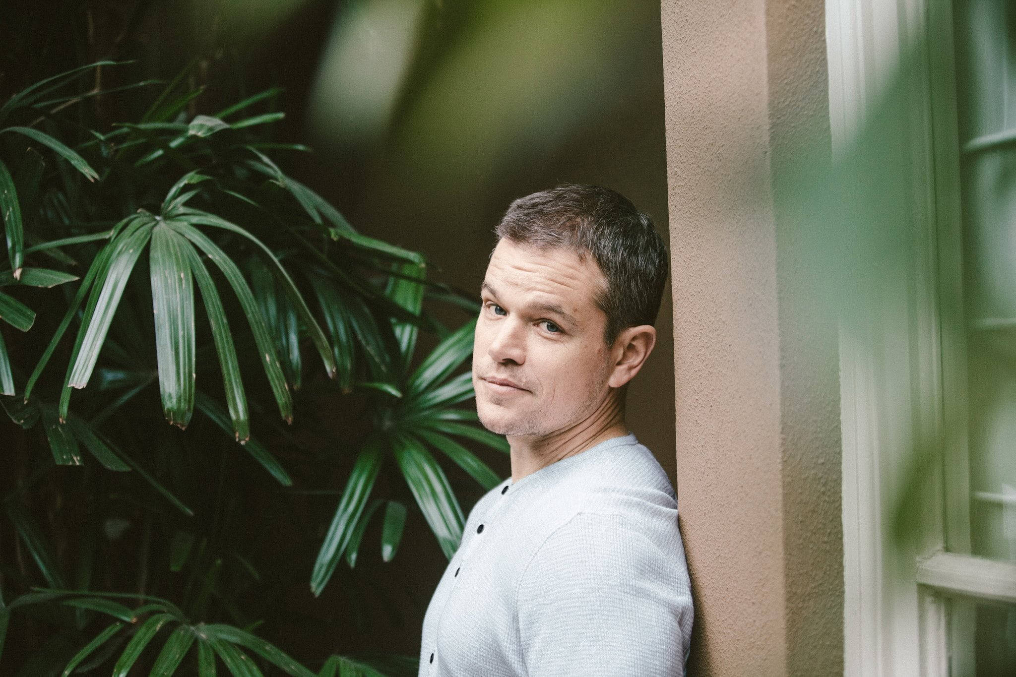 Matt Damon 桌布 壁紙 — 一個男人靠在一面被茂密綠色植物環繞的牆上，光線柔和自然。