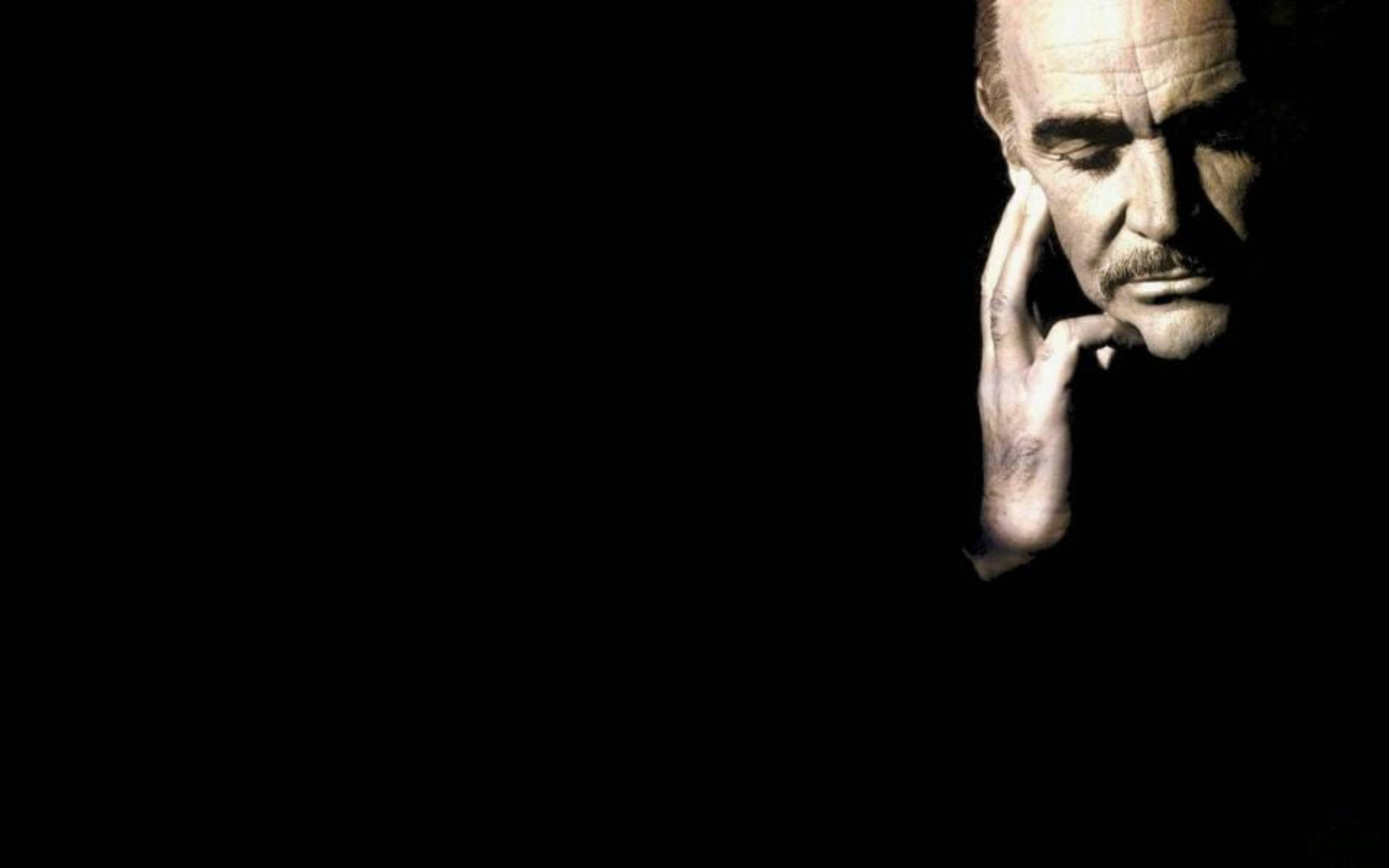 Sean Connery 桌布 壁紙 — 一個男人飽經風霜的臉從黑暗中浮現，手托著臉頰，在戲劇性的聚光燈下擺出沉思的姿勢。