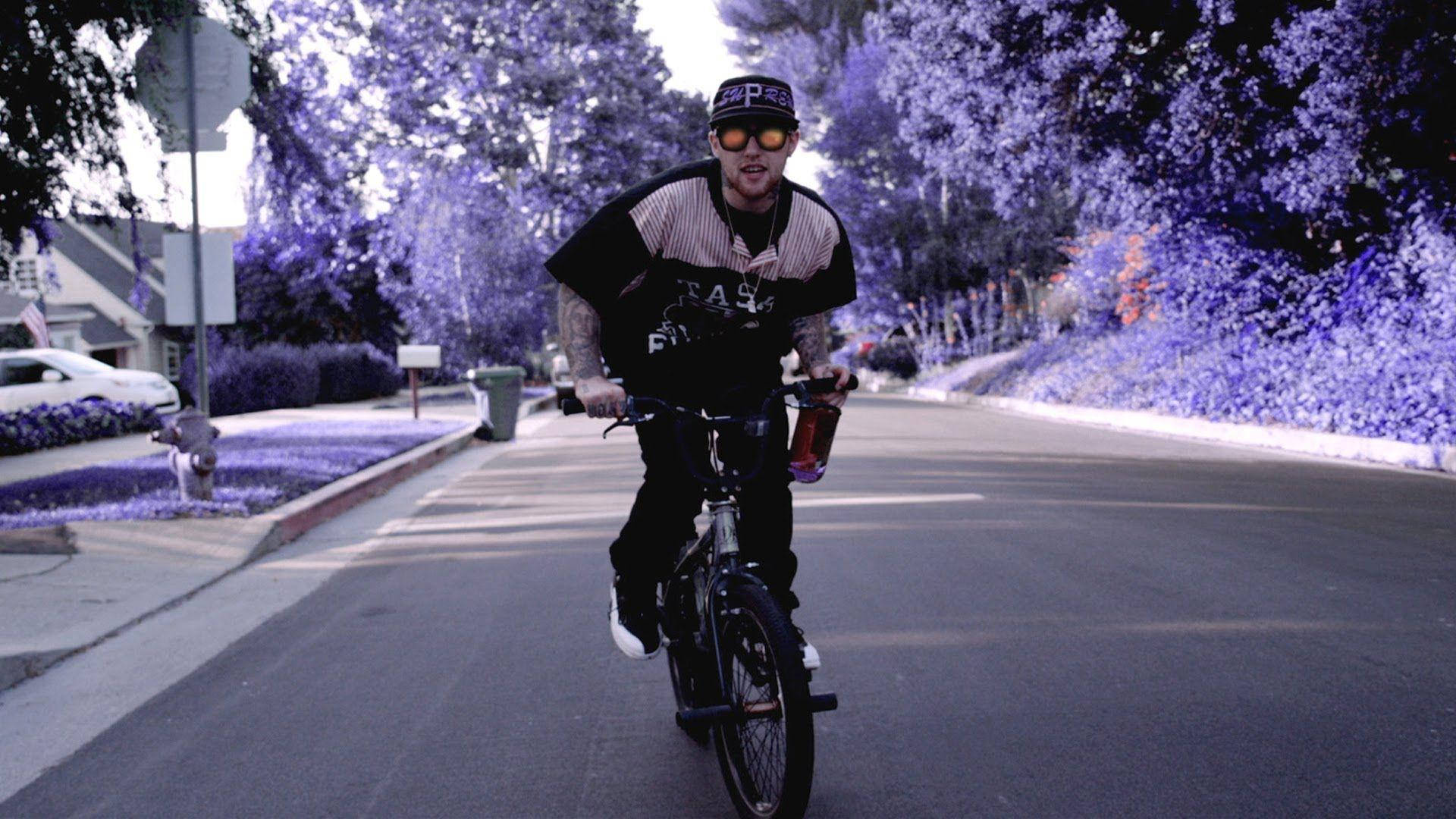 Mac Miller 桌布 壁紙 — 一個男人騎著腳踏車沿著郊區街道行駛，在超現實的紫色天空下，樹木以單色調發光。