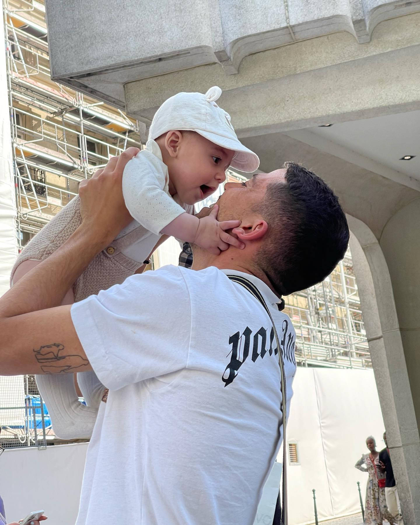 Family 桌布 壁紙 — 一個男人高興地舉起一個戴著白色帽子的嬰兒，他們在混凝土拱門下分享溫馨時刻。