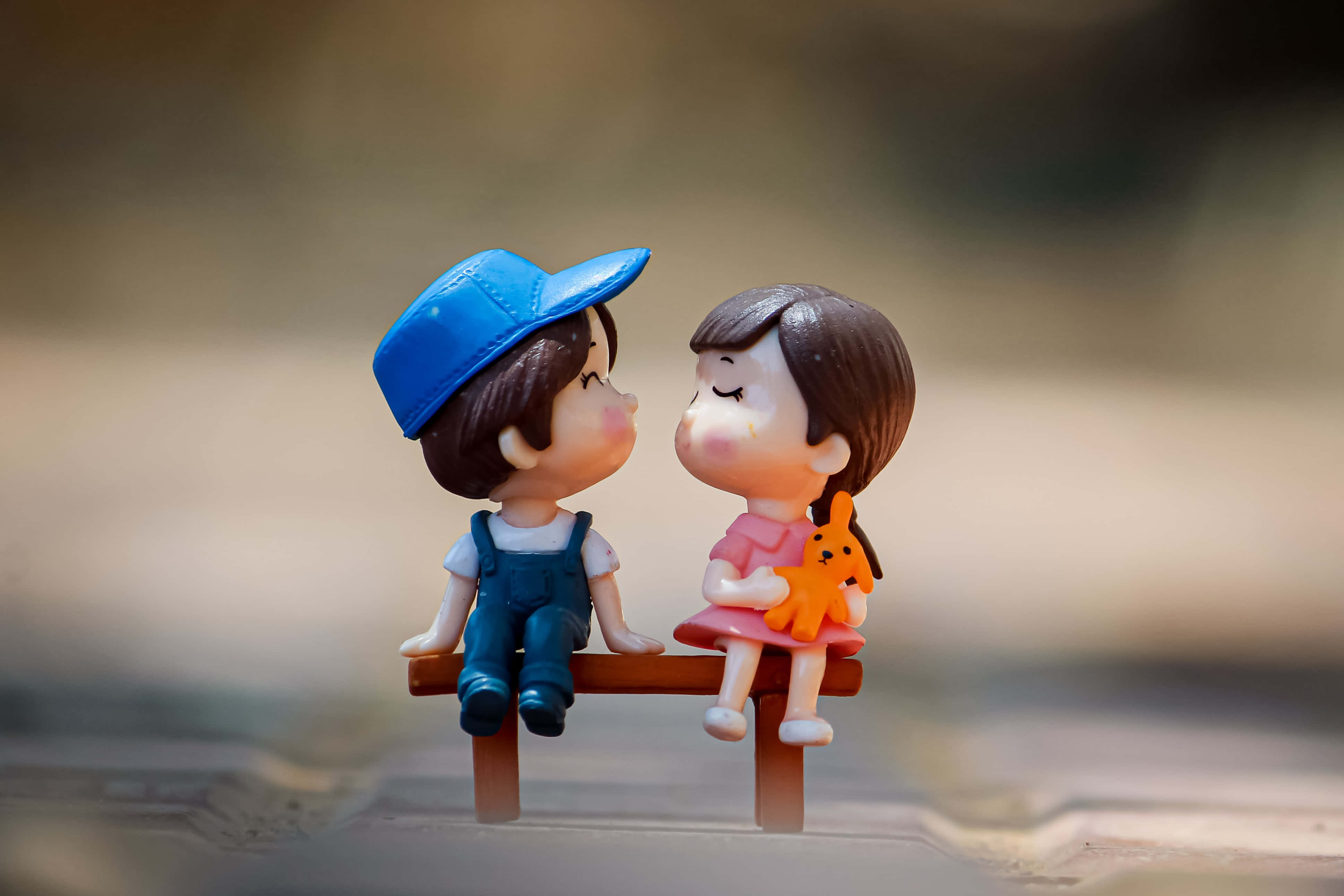 Kiss 桌布 壁紙 — 一個男孩和一個女孩的微型人偶緊挨著坐在長椅上，在柔和自然光下分享溫馨時刻。