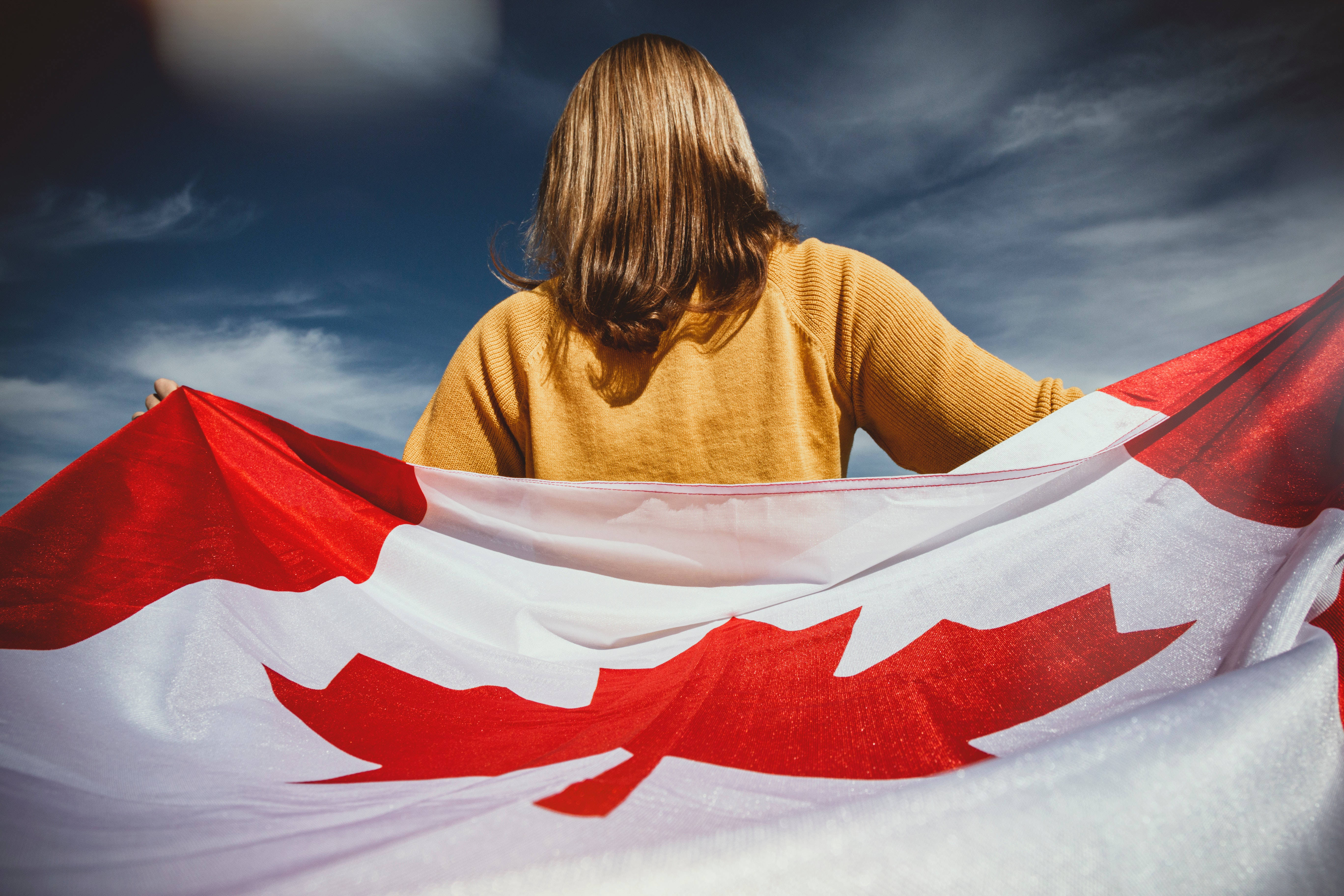 Flag 桌布 壁紙 — 一個留著及肩長髮的人，在戲劇性的多雲天空下舉著一面大型加拿大國旗。