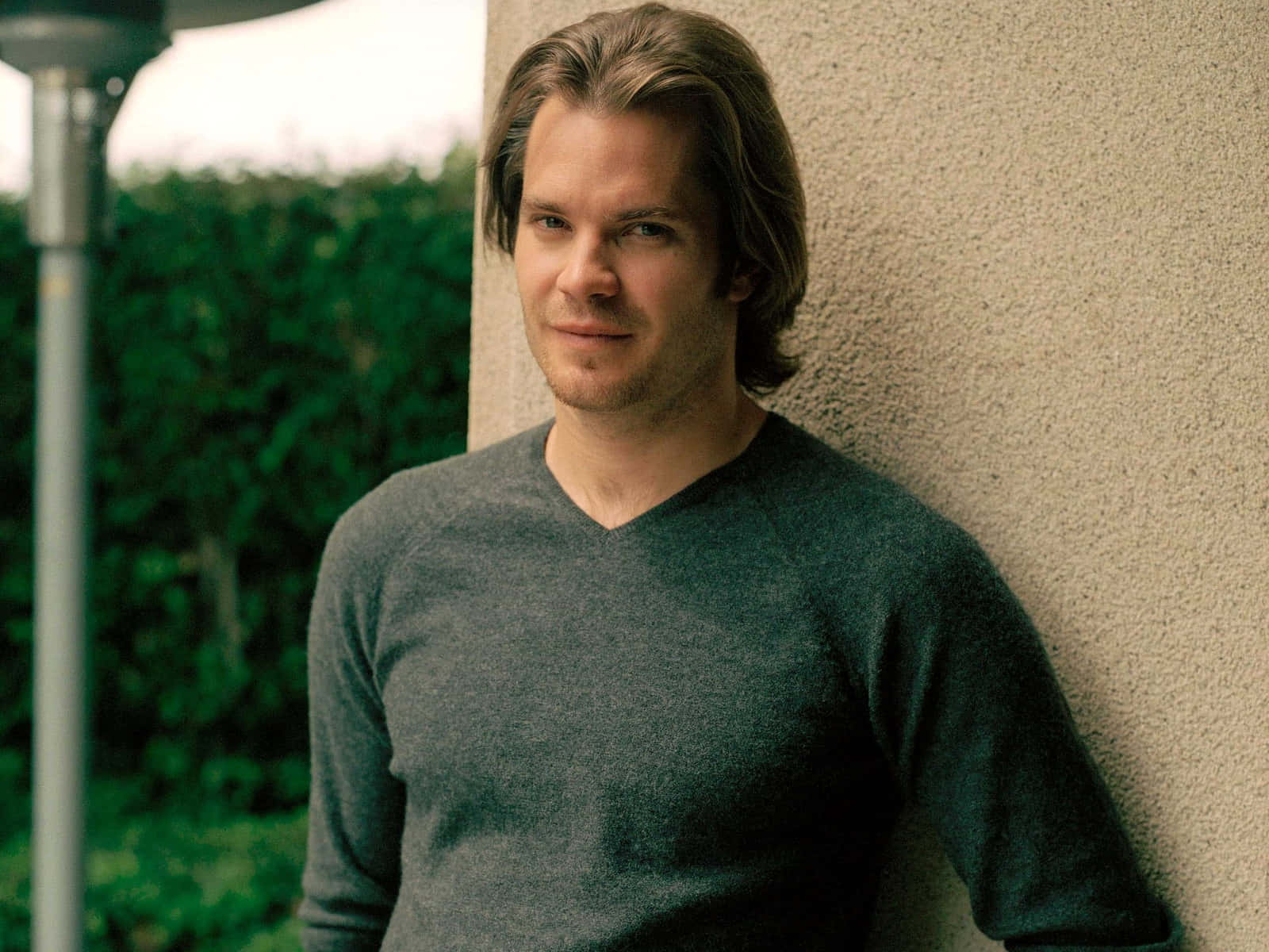 timothy olyphant 桌布 壁紙 — 一個留著及肩長髮的男人，身後有綠色樹葉，隨意地靠在一面有紋理的牆上。