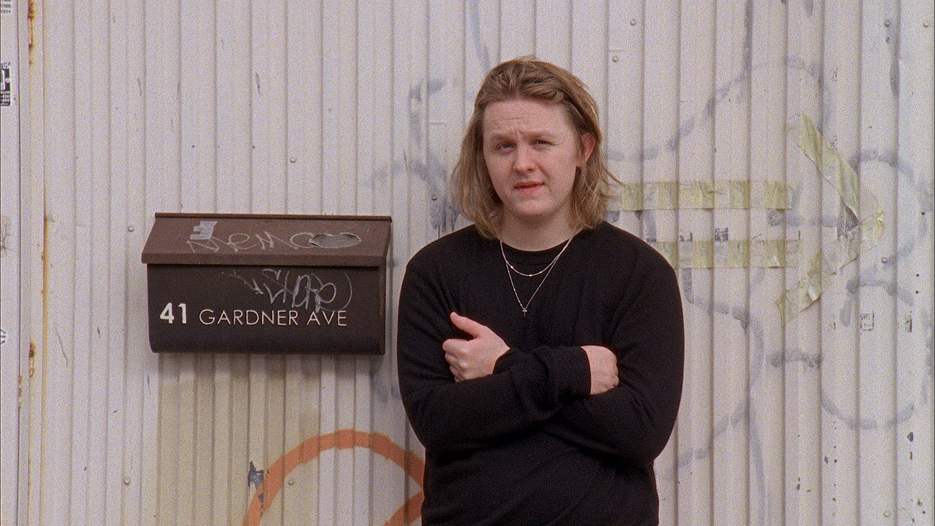 Lewis Capaldi 桌布 壁紙 — 一個留著及肩頭髮的人，雙臂交叉站在一個風化的波紋牆前，牆上有一個被塗鴉標記的郵箱。
