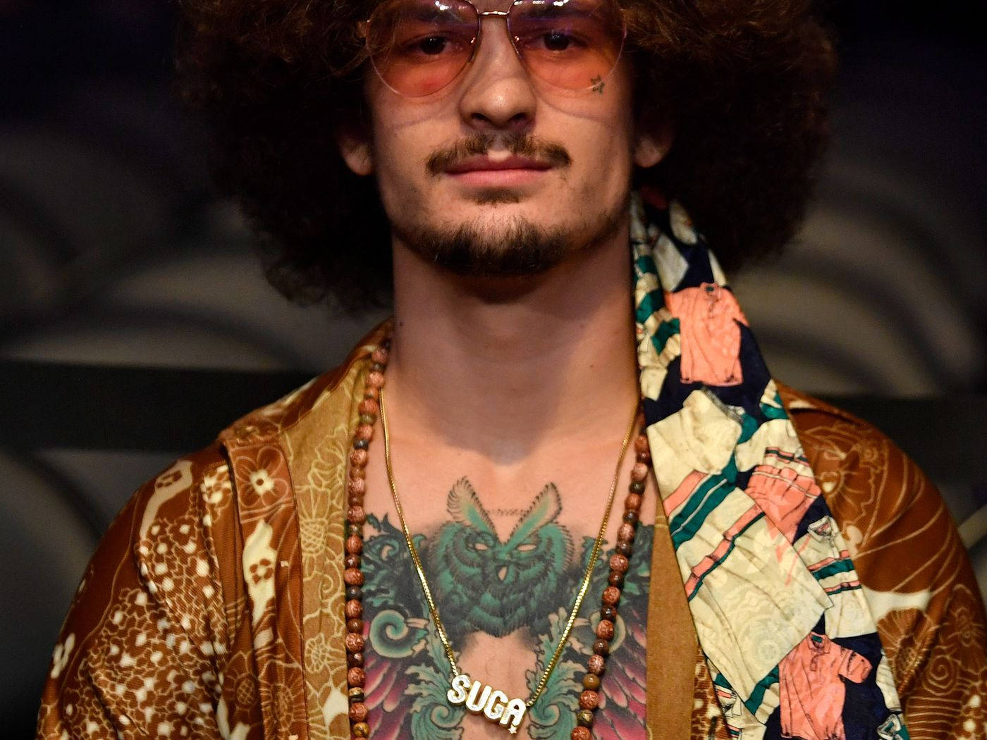 Sunglasses 桌布 壁紙 — 一個留著大爆炸頭、戴著橙色太陽眼鏡的男人，穿著圖案棕色外套和彩色圍巾，展示胸口上的綠色貓頭鷹刺青。