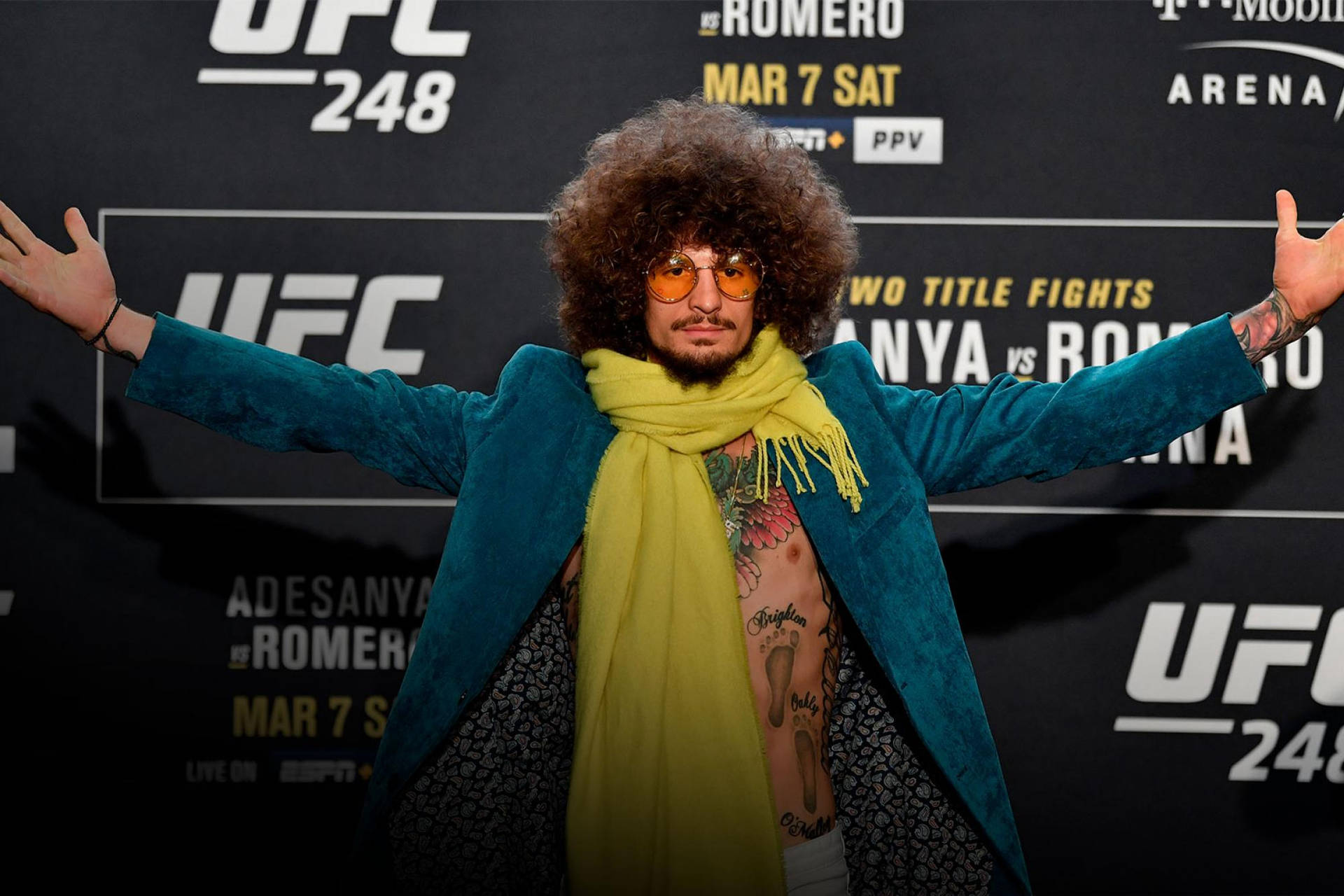 UFC 桌布 壁紙 — 一個留著大爆炸頭、戴著橙色太陽眼鏡的男人，在UFC 248活動背景前張開雙臂，身穿藍綠色外套和黃色圍巾。
