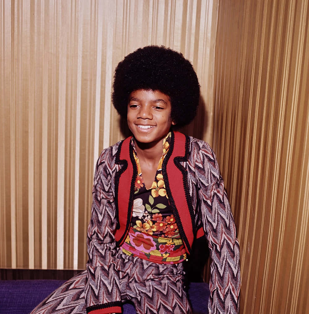 Young Michael Jackson 桌布 壁紙 — 一個留著大爆炸頭的小男孩，穿著色彩繽紛的圖案服裝，在條紋壁紙前燦爛地微笑。