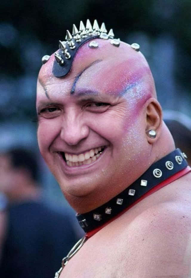 留著尖刺莫霍克頭和閃亮臉部顏料的男人戴著鉚釘項圈微笑。 — 看臉時代 GIF 壁紙