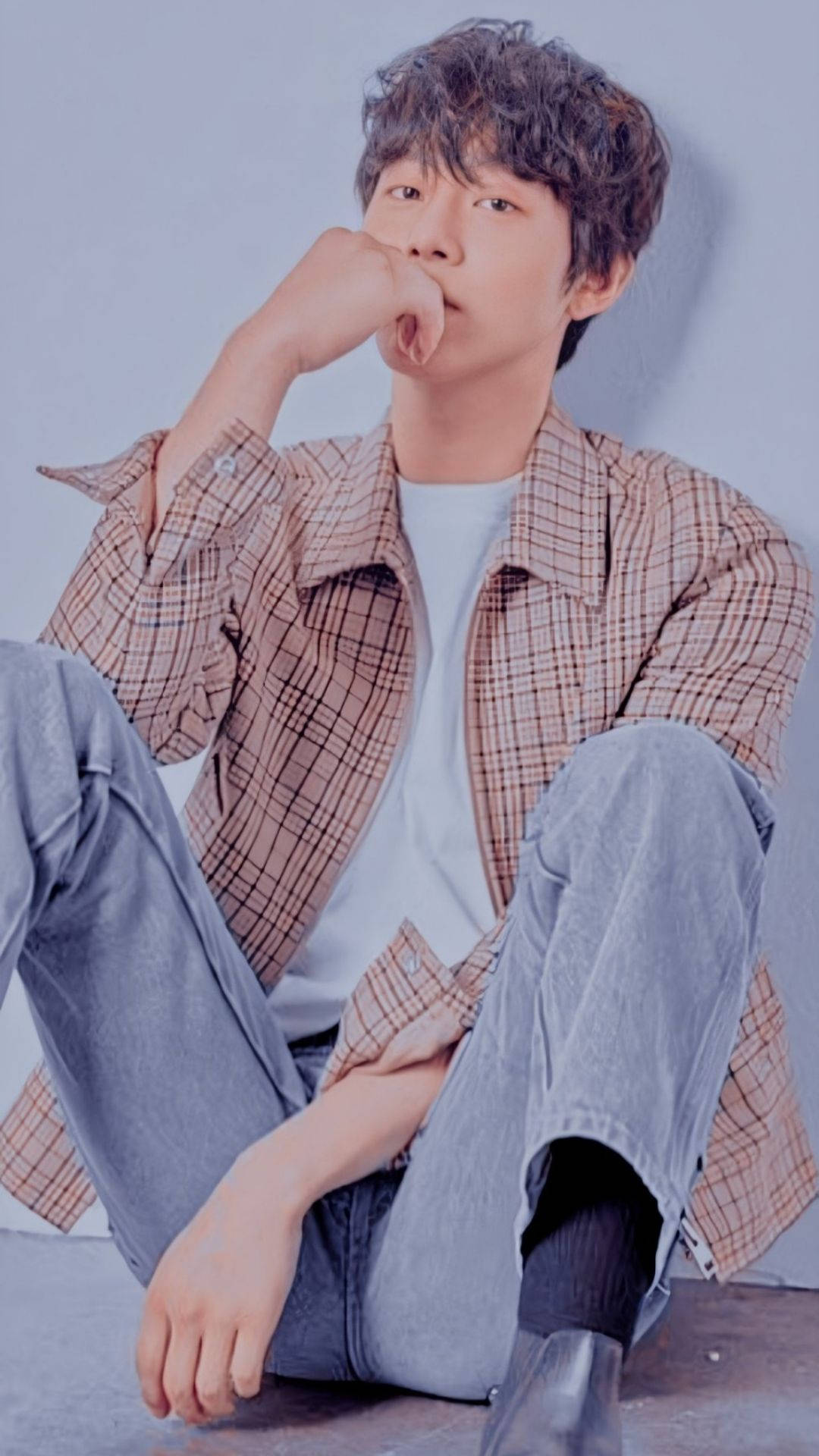 Ahn Hyo Seop 桌布 壁紙 — 一個留著捲髮的年輕男子穿著格紋襯衫和牛仔褲，隨意地坐在地板上，手靠近嘴巴，背景是純藍色的牆壁。