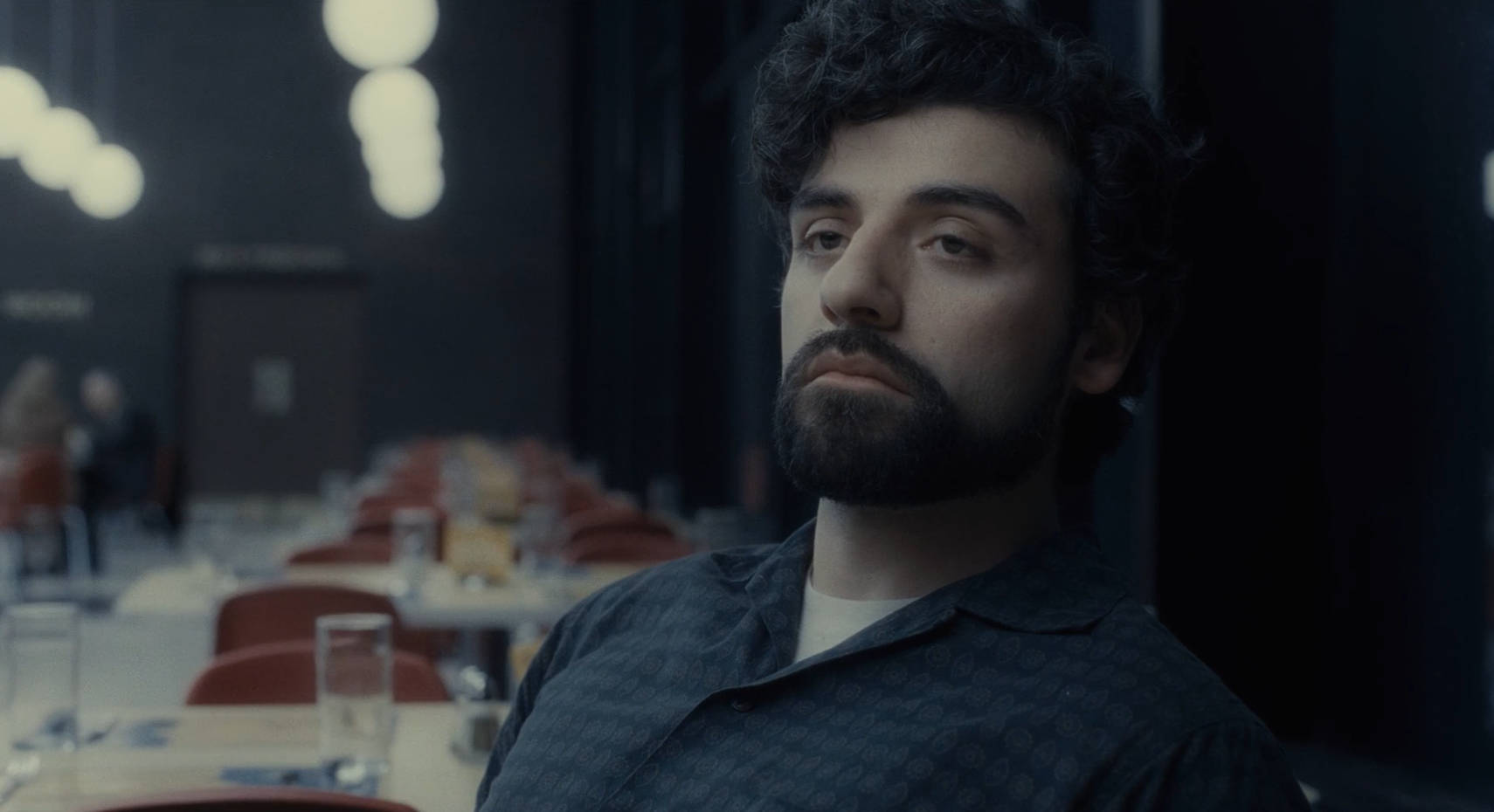 inside llewyn davis 桌布 壁紙 — 一個留著深色捲髮和鬍子的男人，在燈光昏暗、掛著吊燈和空桌的餐廳裡沉思地坐著。