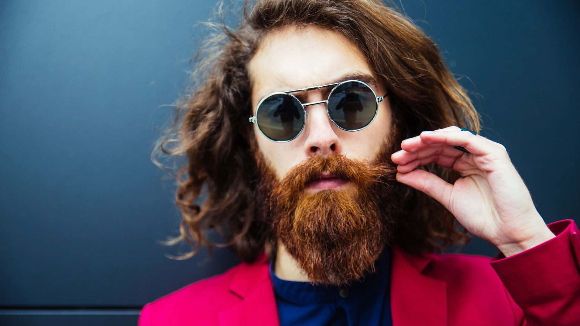 Hipster 桌布 壁紙 — 一個留著濃密紅鬍子、戴著圓形太陽眼鏡的男人，在深藍色牆壁前調整他的眼鏡。
