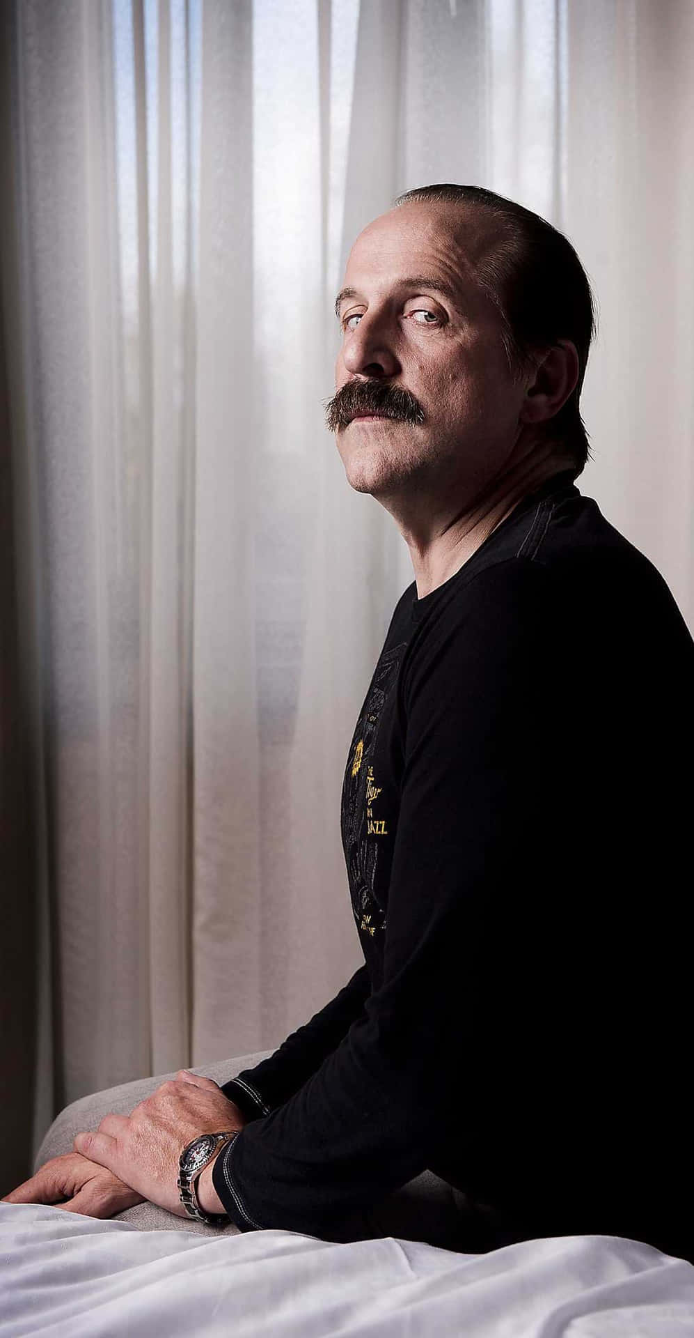 Peter Stormare 桌布 壁紙 — 一個留著濃密鬍子的男人坐在床上，越過肩膀看向鏡頭，柔和的燈光透過他身後的薄窗簾灑進來。