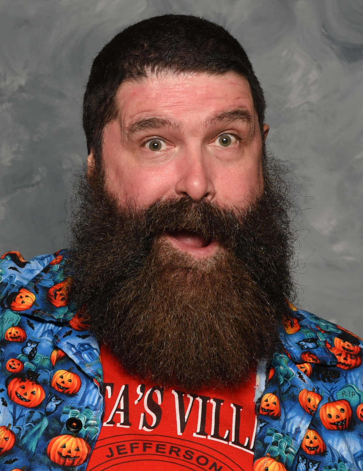 WWE 桌布 壁紙 — 一個留著濃密鬍鬚、眼睛大大的男人，穿著一件印有南瓜燈的萬聖節主題襯衫，背景是灰色攝影棚。